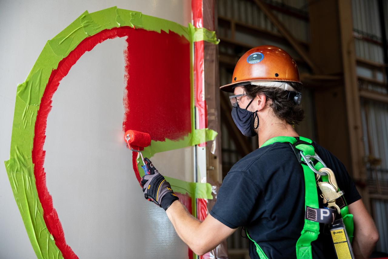 A worker with NASA’s Exploration Ground Systems (EGS) finishes the first coat of the bright red “worm” logo taking shape on the side of an Artemis I solid rocket booster segment inside the Rotation, Processing and Surge Facility (RPSF) at Kennedy Space Center in Florida. The EGS team used a laser projector to mask off the logo with tape, then painted the first coat of the iconic design. The booster segments will help propel the Space Launch System (SLS) rocket on Artemis I, a test of the Orion spacecraft and SLS as an integrated system ahead of crewed flights to the Moon. Northrop Grumman, which built the booster segments, is covering the cost of the painting.