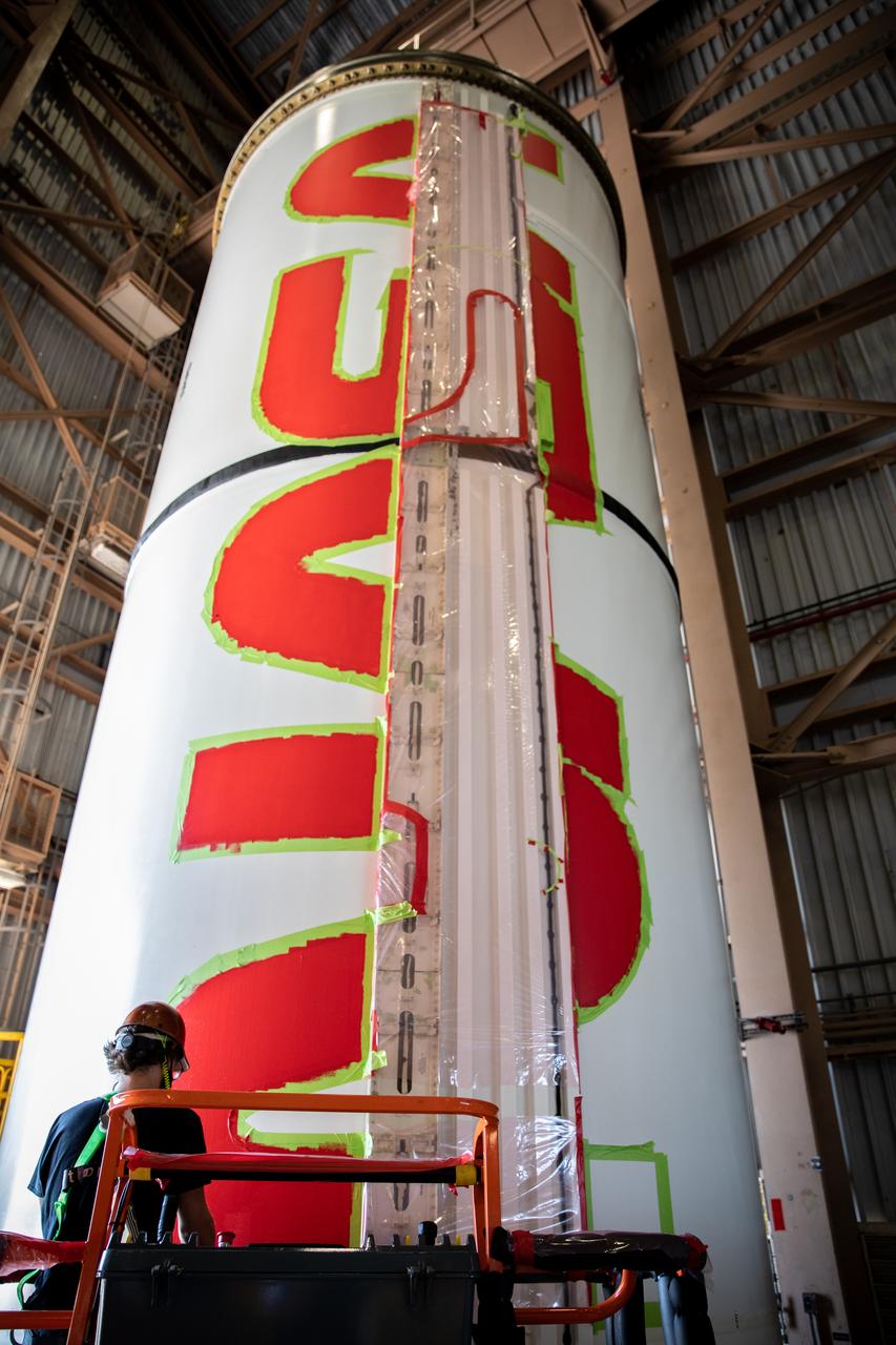 A worker with NASA’s Exploration Ground Systems (EGS) finishes the first coat of the bright red “worm” logo taking shape on the side of an Artemis I solid rocket booster segment inside the Rotation, Processing and Surge Facility (RPSF) at Kennedy Space Center in Florida. The EGS team used a laser projector to mask off the logo with tape, then painted the first coat of the iconic design. The booster segments will help propel the Space Launch System (SLS) rocket on Artemis I, a test of the Orion spacecraft and SLS as an integrated system ahead of crewed flights to the Moon. Northrop Grumman, which built the booster segments, is covering the cost of the painting.