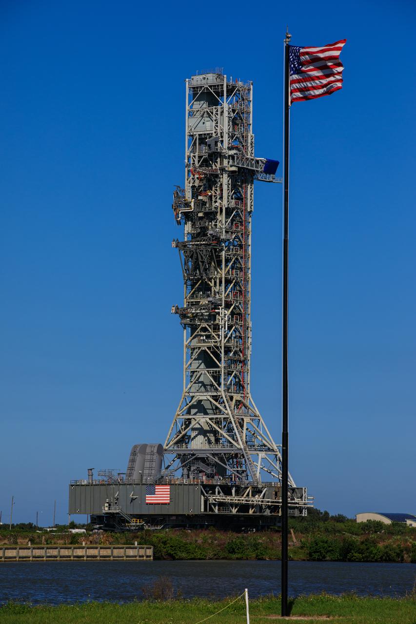 During the morning on Oct. 30, 2020, the mobile launcher for the Artemis I mission, atop crawler-transporter 2, moves slowly along the crawlerway after departing Launch Pad 39B at NASA’s Kennedy Space Center in Florida. In view is the American Flag. The nearly 400-foot-tall mobile launcher is returning to the Vehicle Assembly Building after being at the pad for 10 days, while engineers with Exploration Ground Systems and Jacobs performed several tasks, including a timing test to validate the launch team’s countdown timeline, and a thorough, top-to-bottom wash down of the mobile launcher to remove any debris remaining from construction and installation of the umbilical arms. Artemis I will test the Orion spacecraft and Space Launch System as an integrated system ahead of crewed flights to the Moon. Under the Artemis program, NASA will land the first woman and the next man on the Moon in 2024.