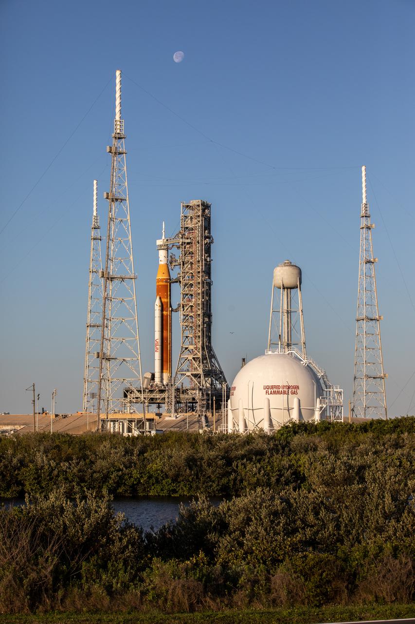 The Moon serves as a background for the Artemis I Space Launch System (SLS) and Orion spacecraft at Launch Pad 39B at NASA’s Kennedy Space Center in Florida on March 21, 2022. The SLS and Orion atop the mobile launcher were transported to the pad on crawler-transporter 2 for a prelaunch test called a wet dress rehearsal. Artemis I will be the first integrated test of the SLS and Orion spacecraft. In later missions, NASA will land the first woman and the first person of color on the surface of the Moon, paving the way for a long-term lunar presence and serving as a steppingstone on the way to Mars. 