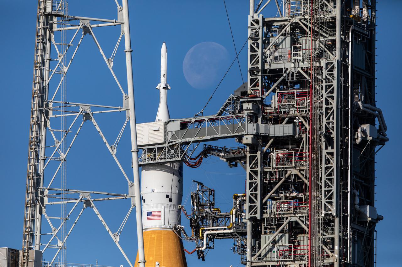 The Moon serves as a background for the Artemis I Space Launch System (SLS) and Orion spacecraft at Launch Pad 39B at NASA’s Kennedy Space Center in Florida on March 21, 2022. The SLS and Orion atop the mobile launcher were transported to the pad on crawler-transporter 2 for a prelaunch test called a wet dress rehearsal. Artemis I will be the first integrated test of the SLS and Orion spacecraft. In later missions, NASA will land the first woman and the first person of color on the surface of the Moon, paving the way for a long-term lunar presence and serving as a steppingstone on the way to Mars. 