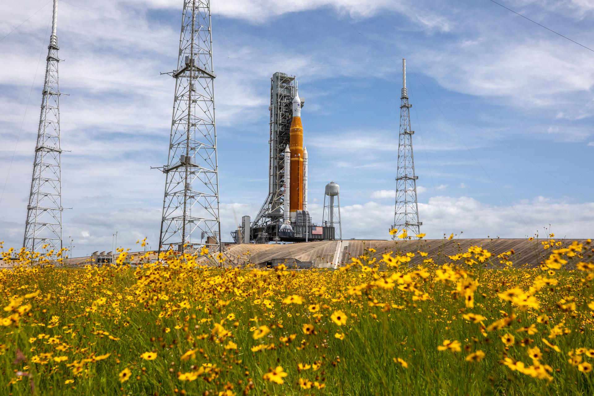 Wildflowers frame a view of the Artemis I Space Launch System (SLS) and Orion spacecraft on Launch Pad 39B at NASA’s Kennedy Space Center in Florida on April 21, 2022. Also in view are the three lightning protection towers and the water tower. The SLS and Orion atop the mobile launcher were transported to the pad on crawler-transporter 2 for a prelaunch test called a wet dress rehearsal. Artemis I will be the first integrated test of the SLS and Orion spacecraft. In future Artemis missions, NASA will land the first woman and the first person of color on the surface of the Moon, paving the way for a long-term lunar presence and serving as a steppingstone on the way to Mars.