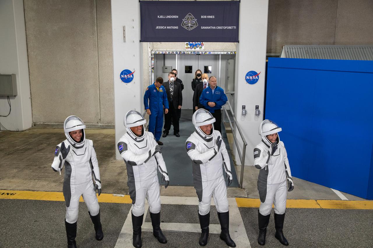 Crew-4 astronauts wave after walking out through the double doors below the Neil A. Armstrong Building’s Astronaut Crew Quarters at NASA’s Kennedy Space Center in Florida on April 27, 2022. They will make their way to the customized Tesla Model X cars that will take them to their spacecraft at Launch Complex 39A. From left are: mission specialist Jessica Watkins, pilot Bob Hines, commander Kjell Lindgren, and mission specialist Samantha Cristoforetti. SpaceX’s Crew Dragon, powered by the company’s Falcon 9 rocket, will carry the four-person crew to the International Space Station as part of NASA’s Commercial Crew Program. Crew-4 is scheduled to launch today at 3:52 a.m. EDT, from Pad 39A at Kennedy.