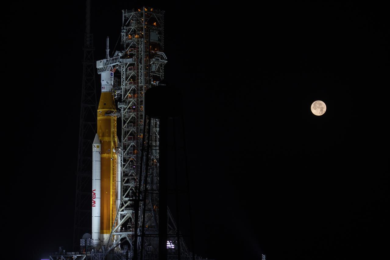 A full Moon is in view from Launch Complex 39B at NASA’s Kennedy Space Center in Florida on June 14, 2022. The Artemis I Space Launch System (SLS) and Orion spacecraft, atop the mobile launcher, are being prepared for a wet dress rehearsal to practice timelines and procedures for launch. The first in an increasingly complex series of missions, Artemis I will test SLS and Orion as an integrated system prior to crewed flights to the Moon. Through Artemis, NASA will land the first woman and first person of color on the lunar surface, paving the way for a long-term lunar presence and using the Moon as a steppingstone on the way to Mars.