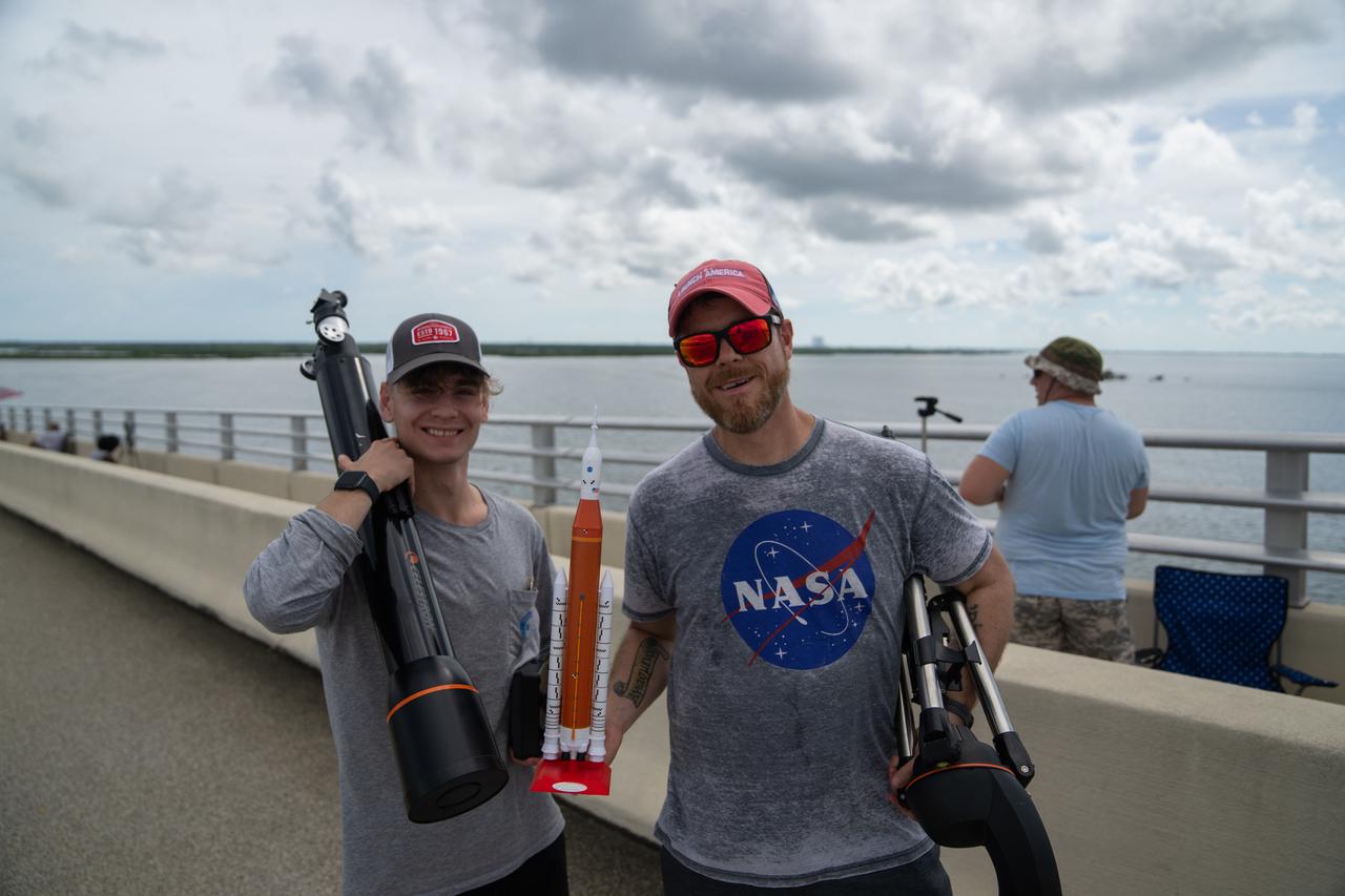 Two individuals wearing NASA shirts and holding a model Space Launch System rocket are on the Max Brewer Bridge in Titusville, Florida, to witness the launch of NASA’s Artemis I mission on Sept. 3, 2022. The launch was waived off for the day. The first in a series of increasingly complex missions, Artemis I will provide a foundation for human deep space exploration and demonstrate our commitment and capability to extend human presence to the Moon and beyond. The primary goal of Artemis I is to thoroughly test the integrated systems before crewed missions by operating the spacecraft in a deep space environment, testing Orion’s heat shield, and recovering the crew module after reentry, descent, and splashdown. 