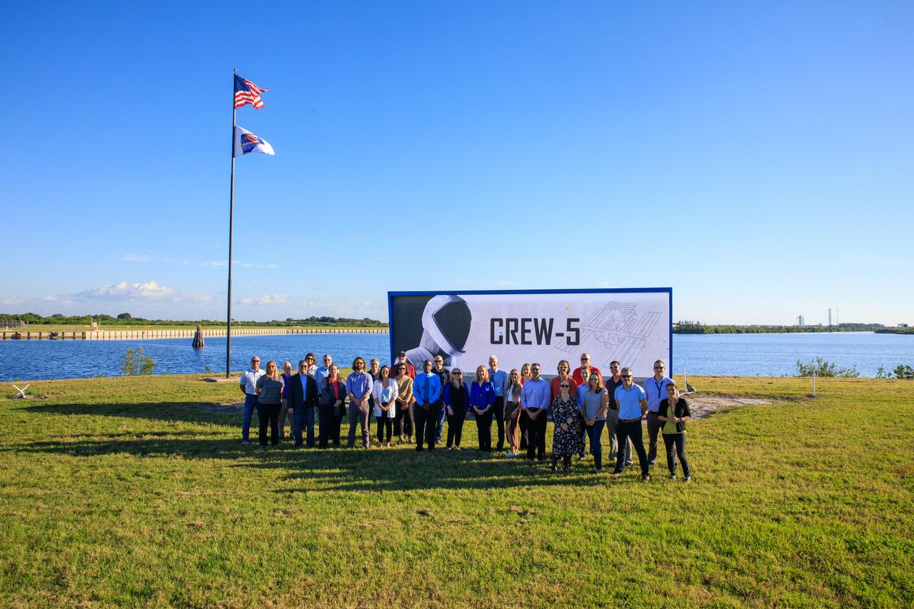 NASA Commercial Crew Program (CCP) employees are photographed in front of Kennedy Space Center’s iconic countdown clock at the Press Site after the Crew-5 flag was raised on Oct. 3, 2022, in Florida. NASA’s SpaceX Crew-5 mission will carry NASA astronauts Josh Cassada and Nicole Aunapu Mann, Roscosmos cosmonaut Anna Kikina, and JAXA (Japan Aerospace Exploration Agency) to the International Space Station for a science expedition mission as part of the agency’s CCP. SpaceX’s Falcon 9 rocket and Crew Dragon Endurance spacecraft are scheduled to lift off from Kennedy’s Launch Complex 39A at noon EDT on Oct. 5, 2022.