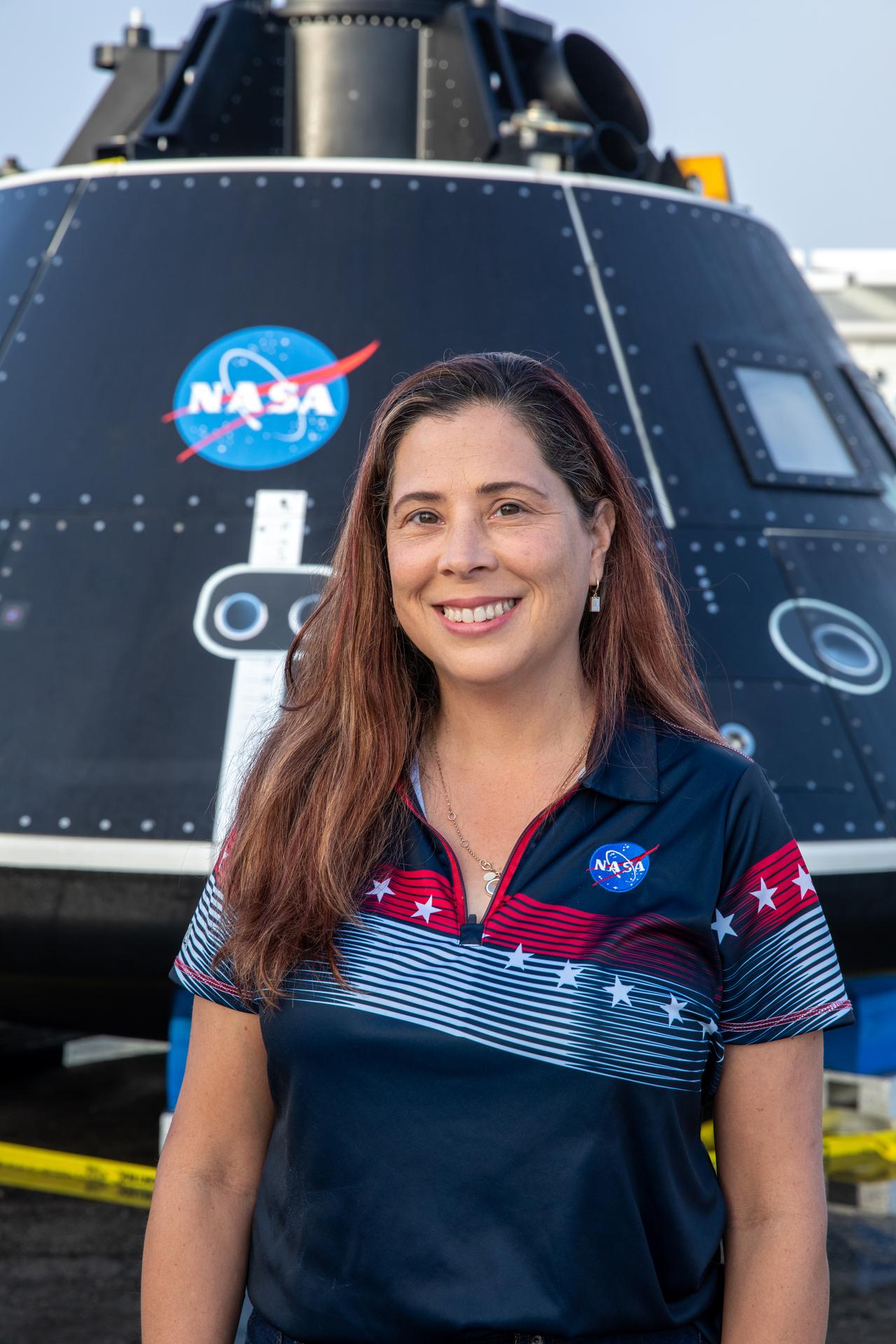 A woman with dark hair, wearing a NASA polo shirt, smiles and poses for a portrait in front of a black spacecraft capsule with the NASA logo.