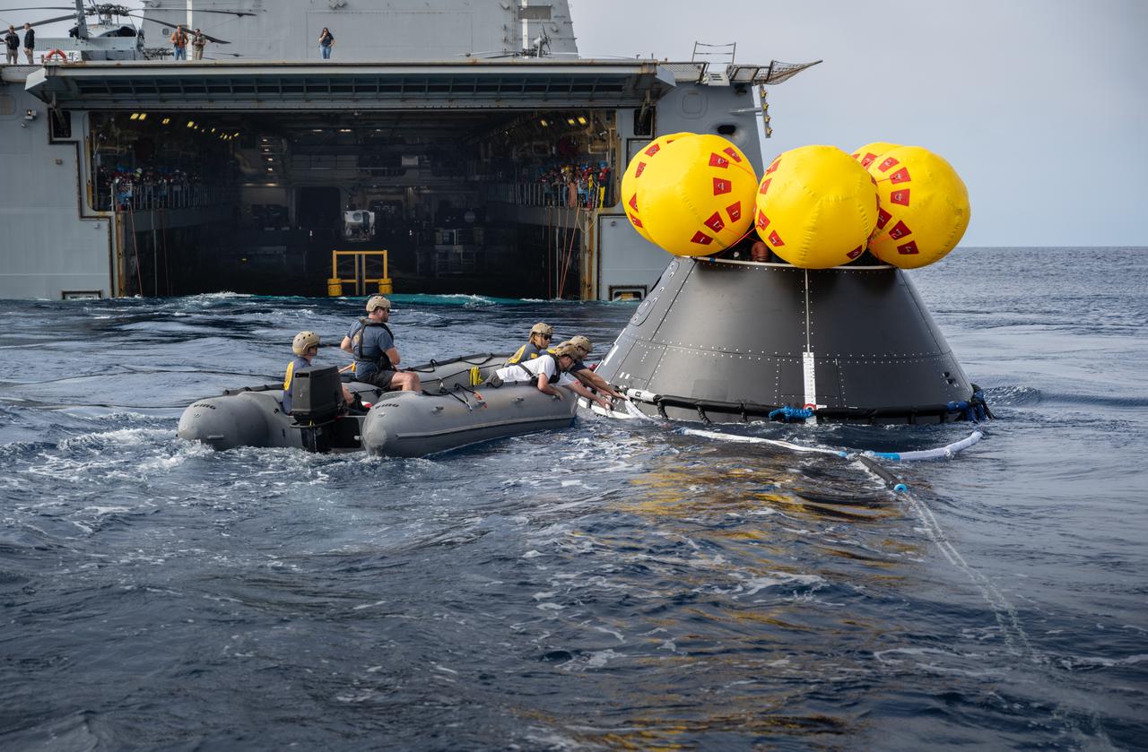 NASA, Navy, and Air Force personnel practice Artemis recovery procedures in the Pacific Ocean as part of Underway Recovery Test 10 (URT-10) off the coast of San Diego. In this photo, Navy divers are seen surrounding the Orion Crew Module Test Article (CMTA) and preparing to recover it inside the recovery ship while simulated astronauts are picked up on an inflatable “front porch” by Naval helicopter pilots from Helicopter Sea Combat Squadron (HSC) 23 “Wildcards” and flown back to the USS John P. Murtha.
