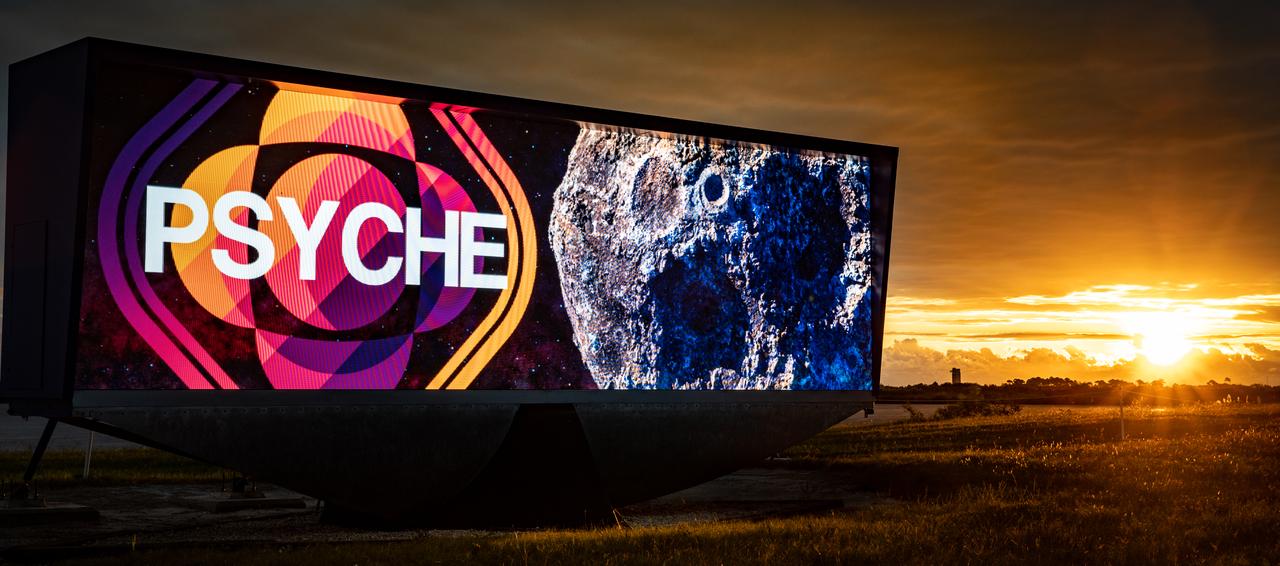 A Psyche mission graphic is displayed on the historic countdown clock as the Sun rises at Kennedy Space Center’s NASA News Center in Florida on Monday, Oct. 16, 2023. NASA’s Psyche spacecraft, atop a SpaceX Falcon Heavy rocket, soared into the sky after lifting off from Kennedy’s Launch Complex 39A at 10:19 a.m. EDT Friday, Oct. 13. The Psyche mission will study a metal-rich asteroid with the same name, located in the main asteroid belt between Mars and Jupiter. This is NASA’s first mission to study an asteroid that has more metal than rock or ice. Riding with Psyche is a pioneering technology demonstration – NASA’s Deep Space Optical Communications (DSOC) experiment – which will be the first test of laser communications beyond the Moon.