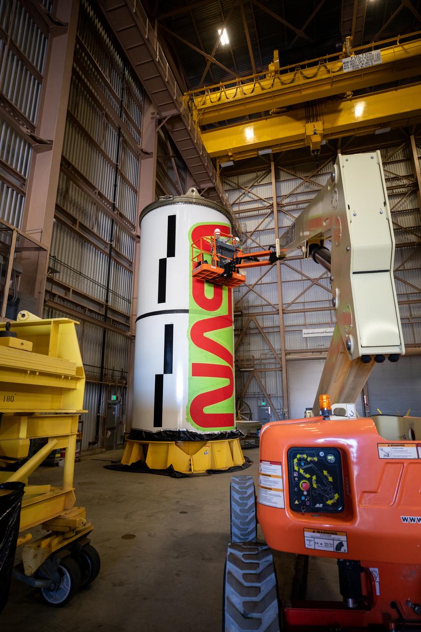 Workers with NASA’s Exploration Ground Systems (EGS) paint the bright red NASA “worm” logo on the side of an Artemis II solid rocket booster segment inside the Rotation, Processing and Surge Facility (RPSF) at Kennedy Space Center in Florida on Tuesday, Jan. 30, 2024. The EGS team used a laser projector to mask off the logo with tape, then painted the first coat of the iconic design. The booster segments will help propel the Space Launch System (SLS) rocket on the Artemis II mission to send four astronauts around the Moon as part of the agency’s effort to establish a long-term science and exploration presence at the Moon, and eventually Mars.