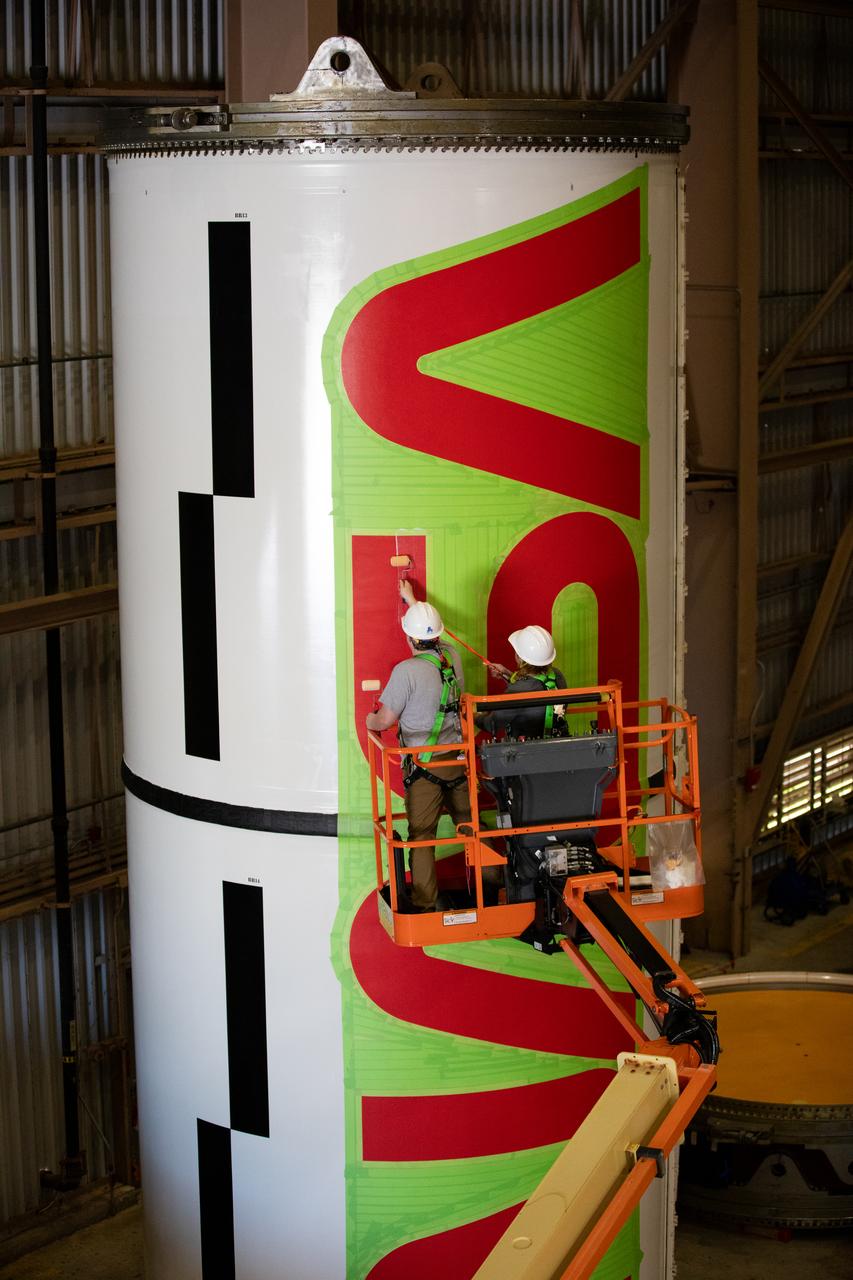 Workers with NASA’s Exploration Ground Systems (EGS) paint the bright red NASA “worm” logo on the side of an Artemis II solid rocket booster segment inside the Rotation, Processing and Surge Facility (RPSF) at Kennedy Space Center in Florida on Tuesday, Jan. 30, 2024. The EGS team used a laser projector to mask off the logo with tape, then painted the first coat of the iconic design. The booster segments will help propel the Space Launch System (SLS) rocket on the Artemis II mission to send four astronauts around the Moon as part of the agency’s effort to establish a long-term science and exploration presence at the Moon, and eventually Mars.