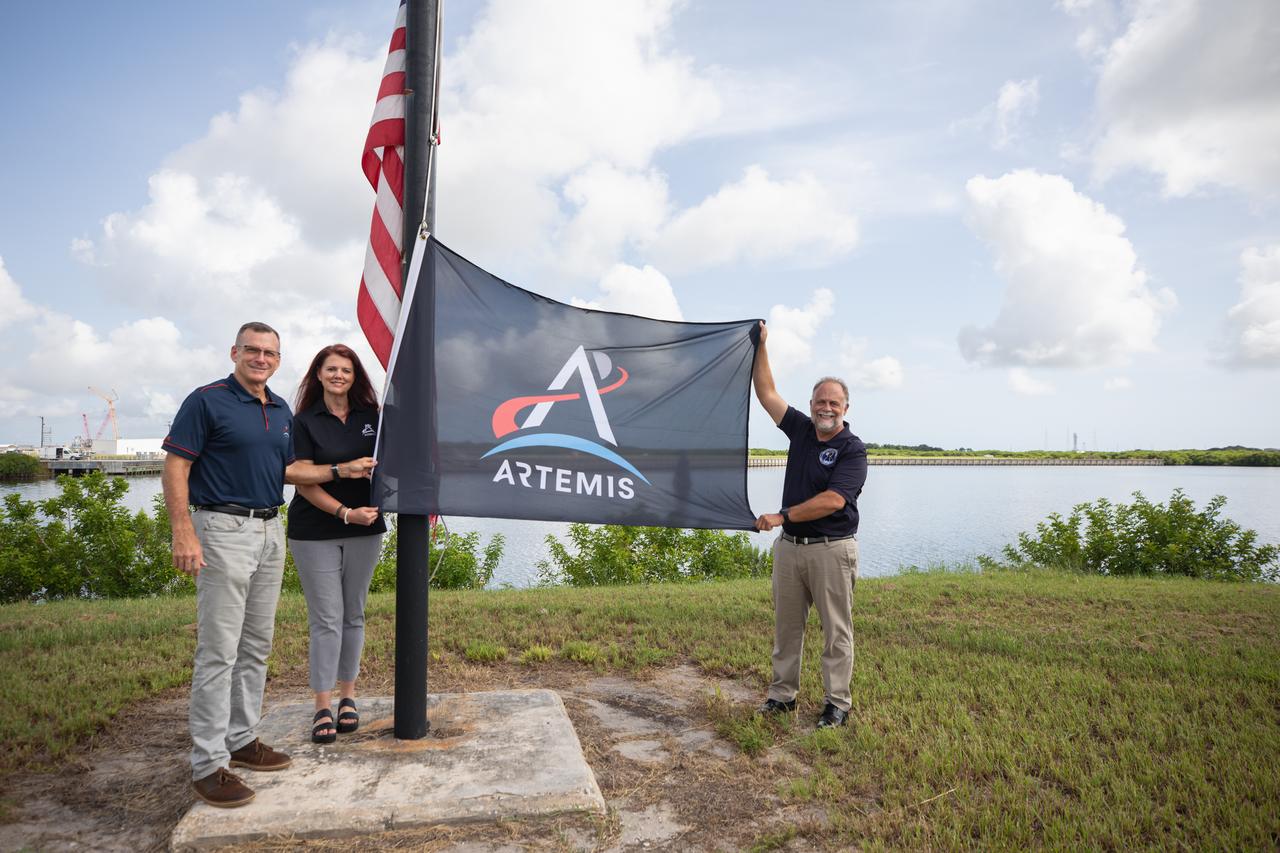 From left to right, Cliff Lanham, senior vehicle operations manager, Exploration Ground Systems; Charlie Blackwell-Thompson, Artemis launch director, Exploration Ground Systems; and Shawn Quinn, manager, Exploration Ground Systems, prepare to raise the Artemis flag near the countdown clock at NASA’s Kennedy Space Center in Florida on Monday, July 22, 2024. The flag raising marks the arrival of NASA’s massive SLS (Space Launch System) core stage via the agency’s Pegasus barge from NASA’s Michoud Assembly Facility in New Orleans. The core stage will help power SLS when it launches four astronauts around the Moon for the first crewed flight of SLS and the Orion spacecraft during the Artemis II test flight.