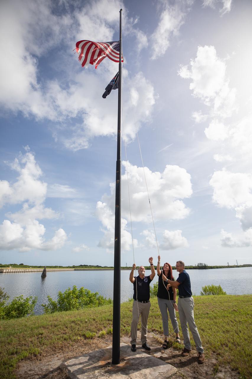 From left to right, Shawn Quinn, manager, Exploration Ground Systems; Charlie Blackwell-Thompson, Artemis launch director, Exploration Ground Systems; and Cliff Lanham, senior vehicle operations manager, Exploration Ground Systems, raise the Artemis flag near the countdown clock at NASA’s Kennedy Space Center in Florida on Monday, July 22, 2024. The flag raising marks the arrival of NASA’s massive SLS (Space Launch System) core stage via the agency’s Pegasus barge from NASA’s Michoud Assembly Facility in New Orleans. The core stage will help power SLS when it launches four astronauts around the Moon for the first crewed flight of SLS and the Orion spacecraft during the Artemis II test flight.
