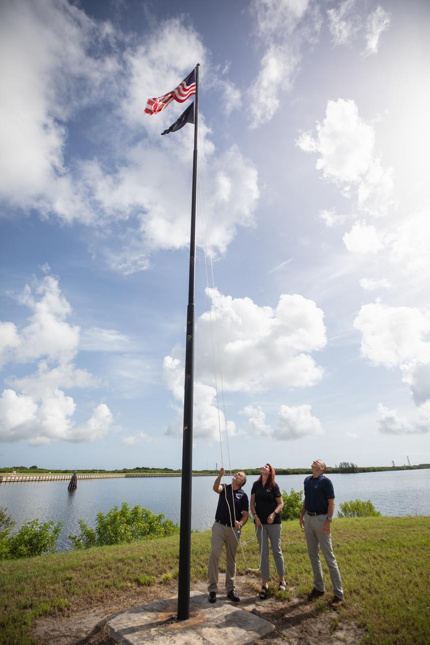 From left to right, Shawn Quinn, manager, Exploration Ground Systems; Charlie Blackwell-Thompson, Artemis launch director, Exploration Ground Systems; and Cliff Lanham, senior vehicle operations manager, Exploration Ground Systems, raise the Artemis flag near the countdown clock at NASA’s Kennedy Space Center in Florida on Monday, July 22, 2024. The flag raising marks the arrival of NASA’s massive SLS (Space Launch System) core stage via the agency’s Pegasus barge from NASA’s Michoud Assembly Facility in New Orleans. The core stage will help power SLS when it launches four astronauts around the Moon for the first crewed flight of SLS and the Orion spacecraft during the Artemis II test flight.