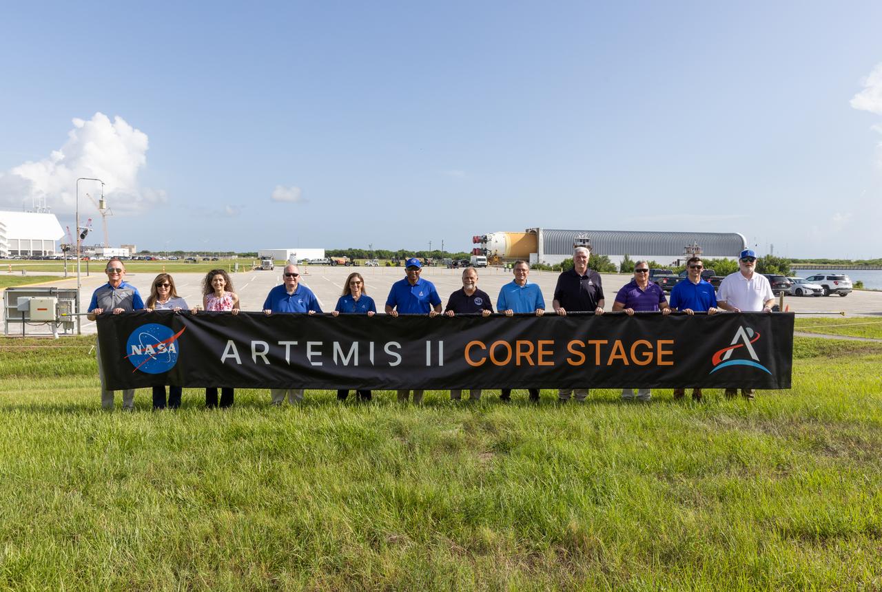 From left, John Ramsey, chief transformation officer for Jacobs; Jennifer Kunz, associate director, technical, NASA’s Kennedy Space Center; Lorna Kenna, vice president and general manager, Jacobs; Burt Summerfield, associate director, management, NASA Kennedy; Catherine Koerner, associate administrator for NASA’s Exploration Systems Development Mission Directorate; Kelvin Manning, deputy director, NASA Kennedy; Shawn Quinn, manager, NASA’s Exploration Ground Systems; Cliff Lanham, senior vehicle operations manager, NASA’s Exploration Ground Systems; Matthew Ramsey, Artemis II mission manager; Paul Hudson, Jacobs senior vehicle engineer; Jay Grow, Boeing’s associate chief engineer for Space Launch System Launch Operations; and Bill Muddle, lead RS-25 field engineer at Aerojet Rocketdyne, hold a banner to celebrate the arrival of the Artemis II SLS (Space Launch System) core stage at NASA’s Kennedy Space Center on Wednesday, July 24, 2024. Journeying from NASA’s Michoud Assembly Facility in New Orleans aboard the Pegasus barge, the core stage arrived at the Florida spaceport on July 23, 2024, to be processed for flight by NASA’s Exploration Ground Systems Program. 