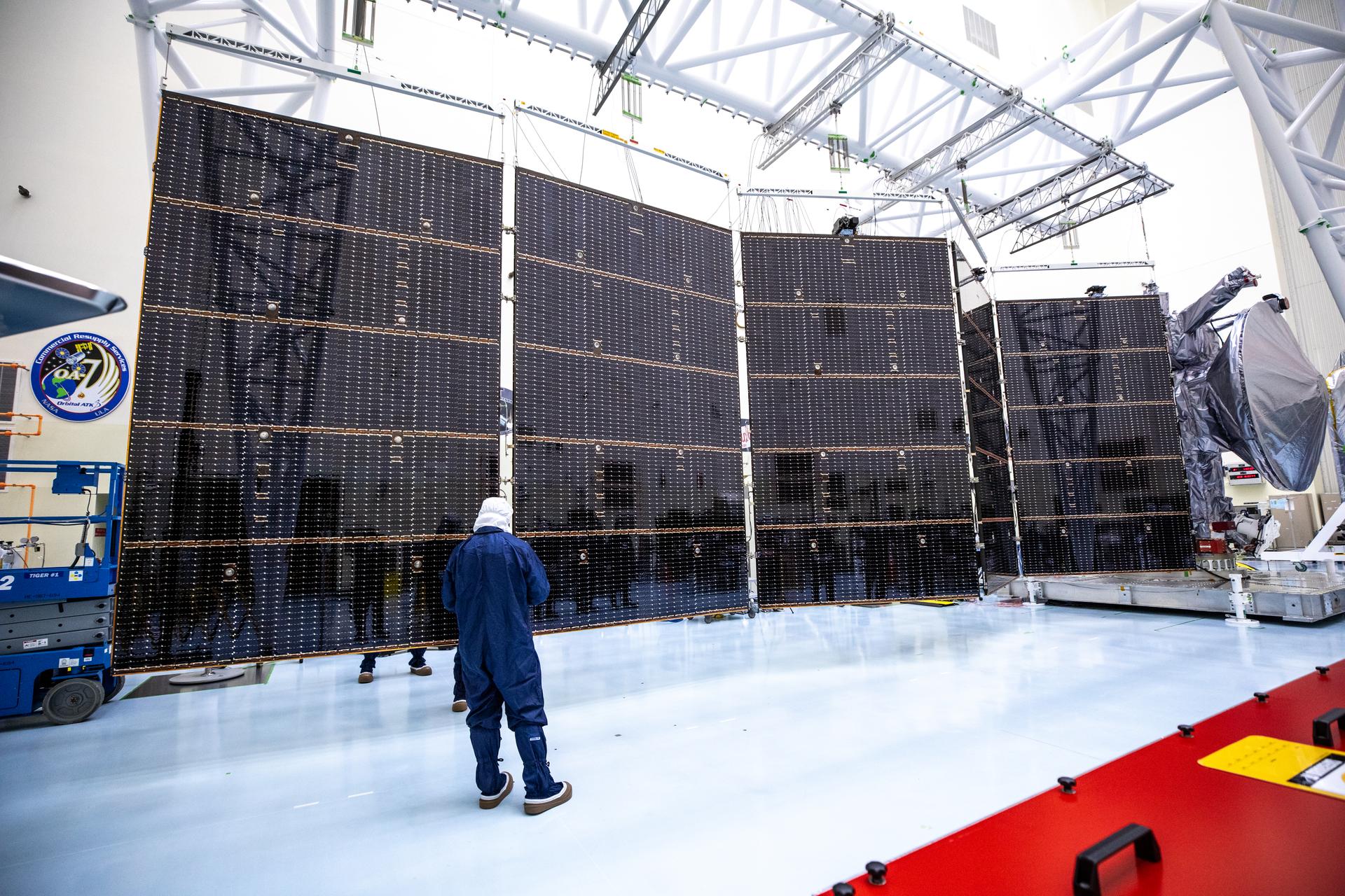 Image shows NASA's Europa Clipper five solar arrays expanded during inspection inside the Payload Hazardous Servicing Facility. On the far right is Europa Clipper's spacecraft wrapped in a silver protective coating ahead of launch. People wearing white and blue protective suits inspect the spacecraft. Photo credit: NASA/Kim Shiflett