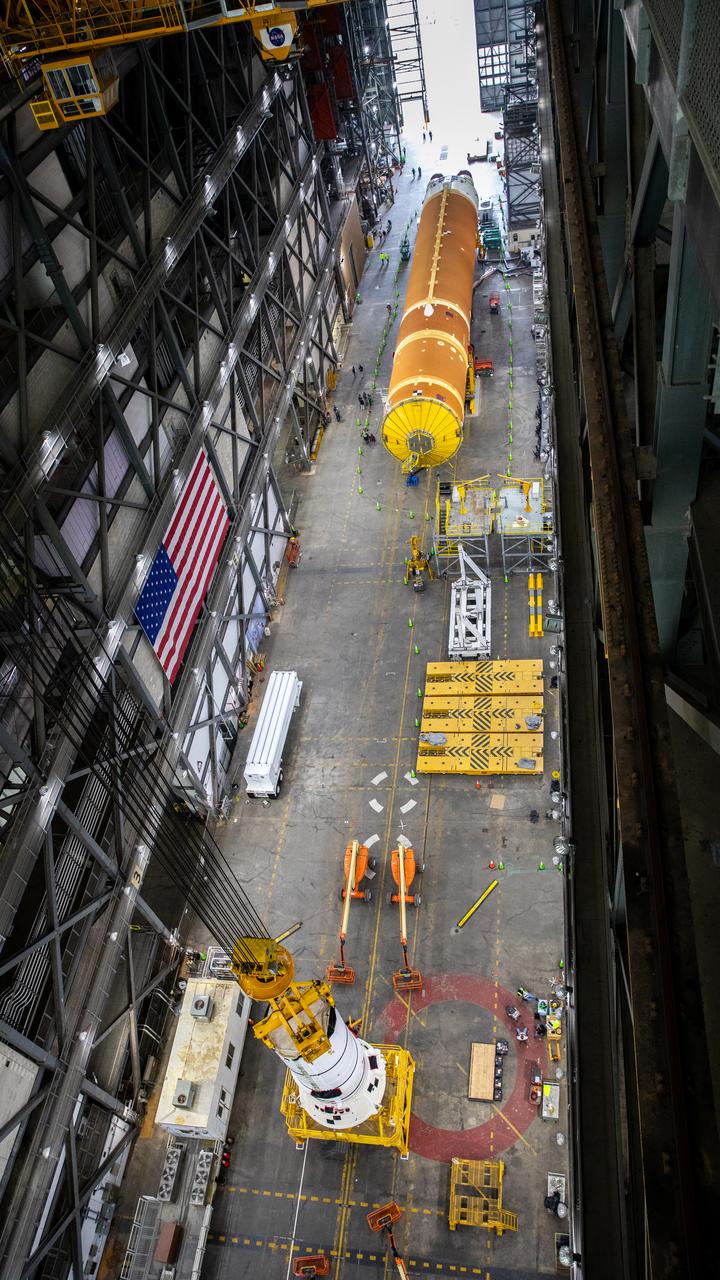 Down the transfer aisle from the Artemis II SLS (Space Launch System) core stage, engineers and technicians with the Exploration Ground Systems Program attach an overhead crane to the left aft assembly, or bottom portion of the solid rocket boosters for the SLS rocket inside the Vehicle Assembly Building at NASA’s Kennedy Space Center on Tuesday, Nov. 19, 2024. The crane will lift the aft assembly on top of the mobile launcher 1 followed by the right aft assembly and stack the remaining booster segments for the Artemis II mission.