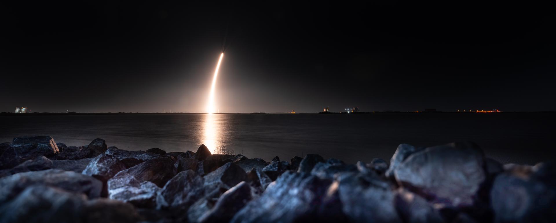 Image shows a night sky with a golden streak reflecting off the water from a launch of a SpaceX Falcon 9 rocket carrying Firefly Aerospace’s Blue Ghost Mission One lander soars upward after liftoff from Launch Complex 39A at NASA’s Kennedy Space Center in Florida on Wednesday, Jan. 15, 2025