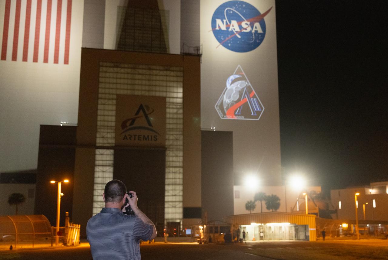 Employees at NASA’s Kennedy Space Center in Florida take photos of the official Artemis II mission crew insignia projected on the exterior of the spaceport’s Vehicle Assembly Building on Friday, April 4, 2025. The patch designates the mission as “AII,” signifying not only the second major flight of the Artemis campaign, but also an endeavor of discovery that seeks to explore for all and by all. Framed in Apollo 8’s famous Earthrise photo, the scene of the Earth and the Moon represents the dual nature of human spaceflight, both equally compelling: The Moon represents our exploration destination, focused on discovery of the unknown. The Earth represents home, focused on the perspective we gain when we look back at our shared planet and learn what it is to be uniquely human. The orbit around Earth highlights the ongoing exploration missions that have enabled Artemis to set sights on a long-term presence on the Moon and soon, Mars.