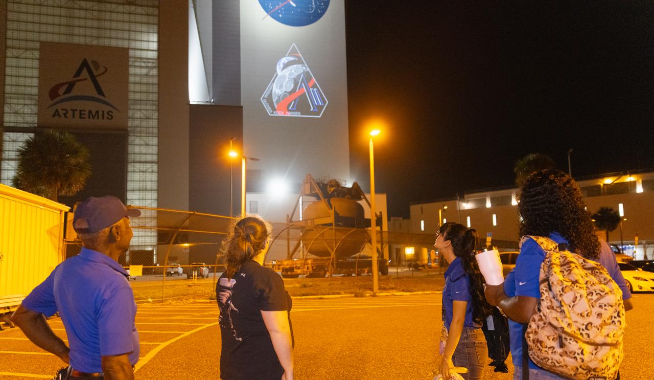Employees at NASA’s Kennedy Space Center in Florida take photos of the official Artemis II mission crew insignia projected on the exterior of the spaceport’s Vehicle Assembly Building on Friday, April 4, 2025. The patch designates the mission as “AII,” signifying not only the second major flight of the Artemis campaign, but also an endeavor of discovery that seeks to explore for all and by all. Framed in Apollo 8’s famous Earthrise photo, the scene of the Earth and the Moon represents the dual nature of human spaceflight, both equally compelling: The Moon represents our exploration destination, focused on discovery of the unknown. The Earth represents home, focused on the perspective we gain when we look back at our shared planet and learn what it is to be uniquely human. The orbit around Earth highlights the ongoing exploration missions that have enabled Artemis to set sights on a long-term presence on the Moon and soon, Mars.