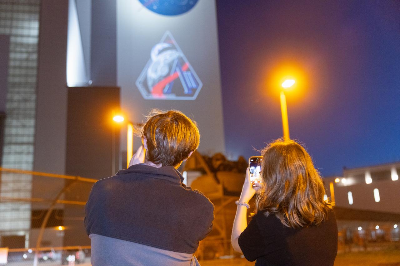 Employees at NASA’s Kennedy Space Center in Florida take photos of the official Artemis II mission crew insignia projected on the exterior of the spaceport’s Vehicle Assembly Building on Friday, April 4, 2025. The patch designates the mission as “AII,” signifying not only the second major flight of the Artemis campaign, but also an endeavor of discovery that seeks to explore for all and by all. Framed in Apollo 8’s famous Earthrise photo, the scene of the Earth and the Moon represents the dual nature of human spaceflight, both equally compelling: The Moon represents our exploration destination, focused on discovery of the unknown. The Earth represents home, focused on the perspective we gain when we look back at our shared planet and learn what it is to be uniquely human. The orbit around Earth highlights the ongoing exploration missions that have enabled Artemis to set sights on a long-term presence on the Moon and soon, Mars.