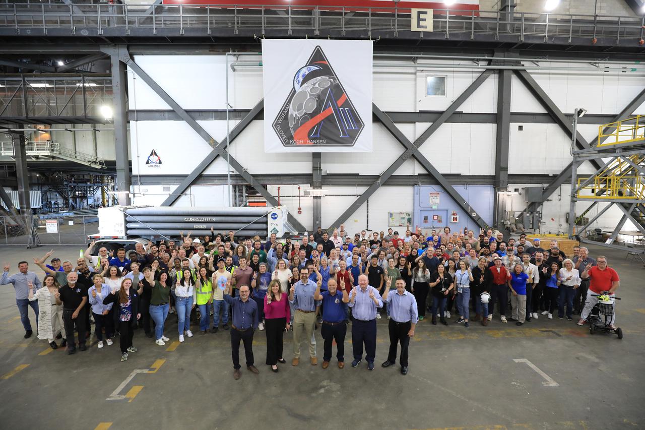 Members of NASA leadership, leadership with the agency’s Exploration Ground Systems Program, and contractor Amentum Services, Inc. leadership pose for a group photo underneath a banner displaying the Artemis II mission insignia outside High Bay 3 at the Vehicle Assembly Building at NASA’s Kennedy Space Center in Florida on Thursday, April 10, 2025. The design designates the mission as “AII,” signifying not only the second major flight of the Artemis campaign, but also an endeavor of discovery that seeks to explore for all and by all. Framed in Apollo 8’s famous Earthrise photo, the scene of the Earth and the Moon represents the dual nature of human spaceflight, both equally compelling: The Moon represents our exploration destination, focused on discovery of the unknown. The Earth represents home, focused on the perspective we gain when we look back at our shared planet and learn what it is to be uniquely human. The orbit around Earth highlights the ongoing exploration missions that have enabled Artemis to set sights on a long-term presence on the Moon and soon, Mars. 