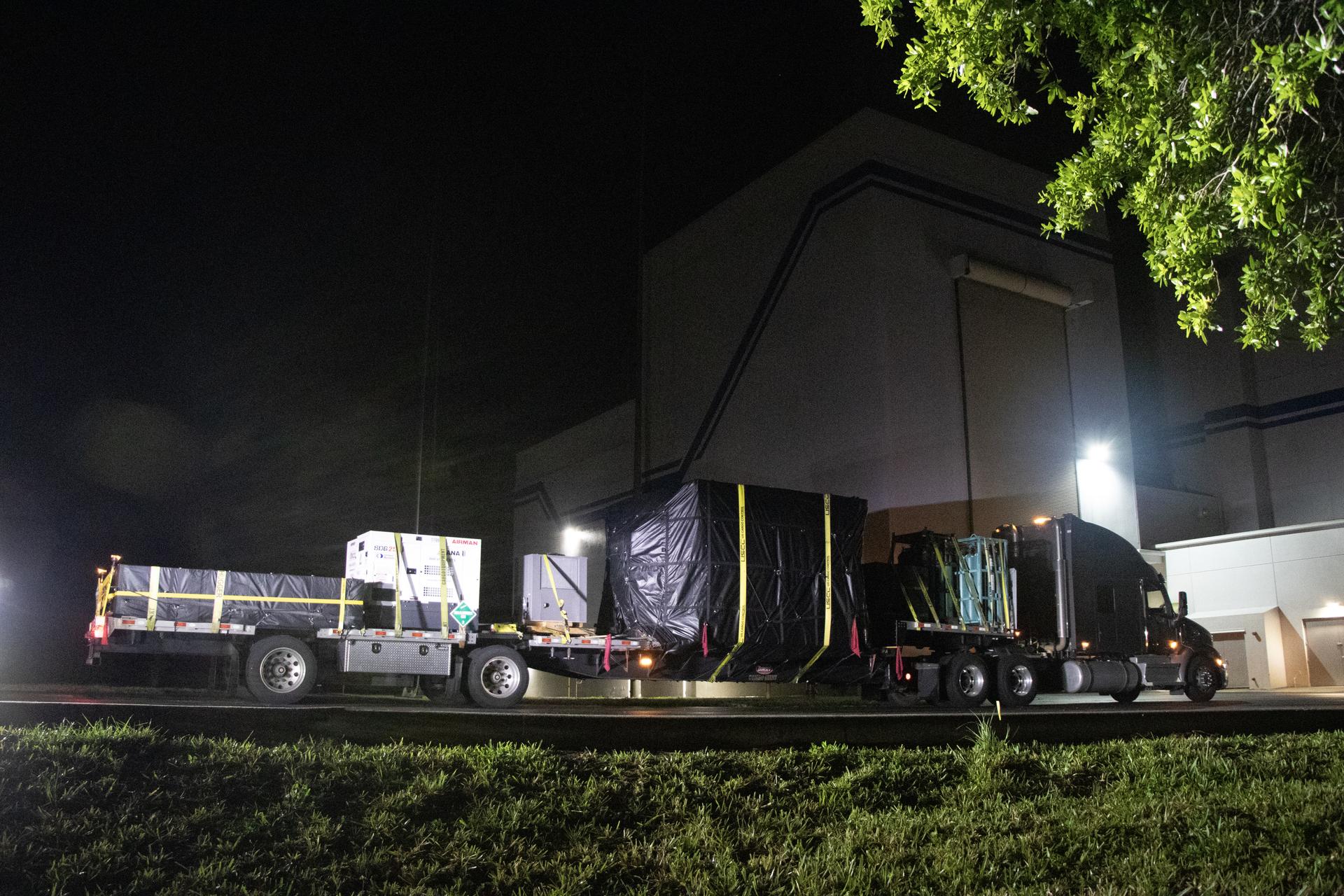 semitractor-trailer pulls a long, multi-axle flatbed trailer carrying a large metal box covered in a tarp that contains NASA's Interstellar Mapping and Acceleration Probe or IMAP spacecraft, along with a white generator and several other other containers. It is front of a tall, white industrial building with a larger roll-up door.