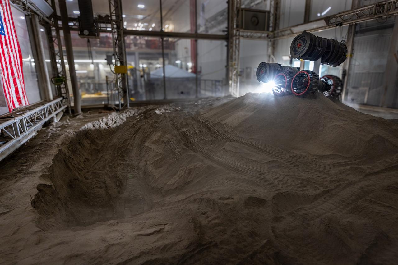 NASA’s RASSOR (Regolith Advanced Surface Systems Operations Robot) manipulates simulated regolith, or lunar dust found on the Moon’s surface, to create a three-foot berm during a site preparation test inside of the Granular Mechanics and Regolith Operations Lab at the agency’s Kennedy Space Center in Florida on Tuesday, June 3, 2025. The opposing motion of the bucket drums helps RASSOR grip the surface in low-gravity environments like the Moon or Mars. With this unique capability, RASSOR can traverse the rough surface to dig, load, haul, and dump regolith that could be used in construction or broken down into hydrogen, oxygen, or water, resources critical for sustaining human presence. RASSOR represents an earlier generation technology that informed the development of NASA’s IPEx (In-Situ Resource Utilization Pilot Excavator), serving as a precursor and foundational platform for the advanced excavation systems and autonomous capabilities now being demonstrated by this Moon-mining robot.