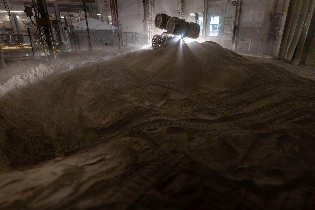 NASA’s RASSOR (Regolith Advanced Surface Systems Operations Robot) manipulates simulated regolith, or lunar dust found on the Moon’s surface, to create a three-foot berm during a site preparation test inside of the Granular Mechanics and Regolith Operations Lab at the agency’s Kennedy Space Center in Florida on Tuesday, June 3, 2025. The opposing motion of the bucket drums helps RASSOR grip the surface in low-gravity environments like the Moon or Mars. With this unique capability, RASSOR can traverse the rough surface to dig, load, haul, and dump regolith that could be used in construction or broken down into hydrogen, oxygen, or water, resources critical for sustaining human presence. RASSOR represents an earlier generation technology that informed the development of NASA’s IPEx (In-Situ Resource Utilization Pilot Excavator), serving as a precursor and foundational platform for the advanced excavation systems and autonomous capabilities now being demonstrated by this Moon-mining robot.