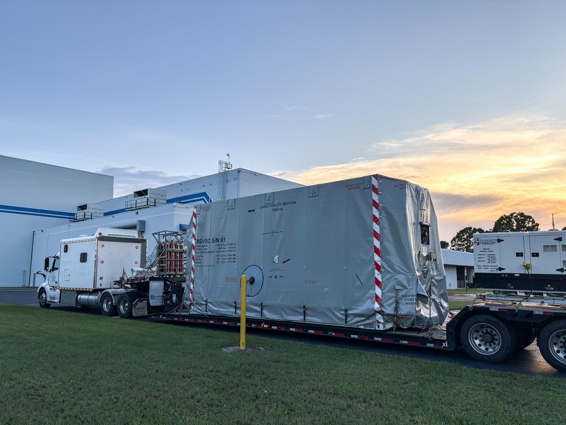 Image shows a semi-trailer carrying a satellite covered in a silver protective cover at Astrotech Space Operations near NASA's Kennedy Space Center.