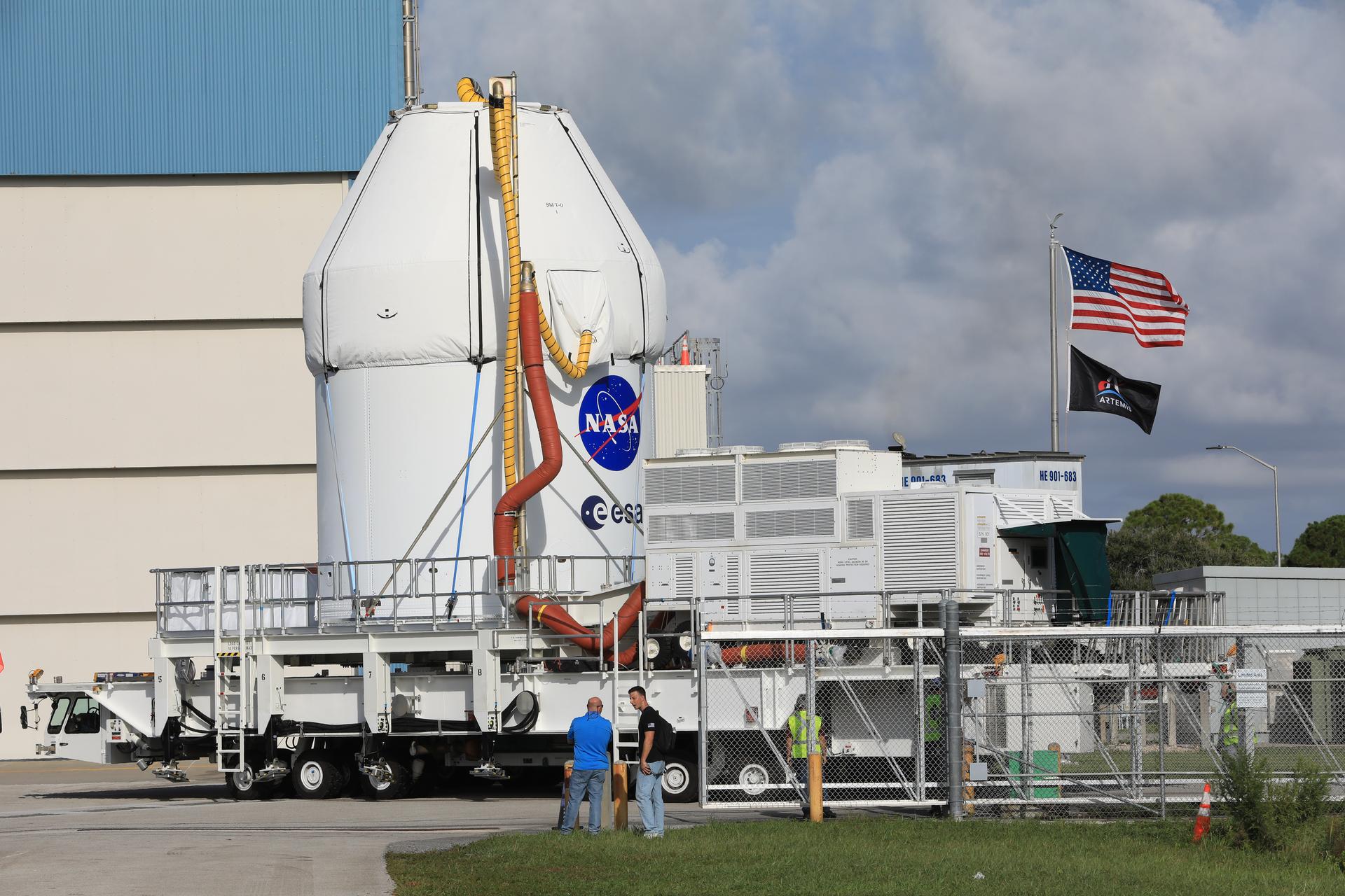 Image shows a white piece of hardware with the NASA insignia transported across NASA Kennedy. An American flag and Artemis flag are seen in the background.