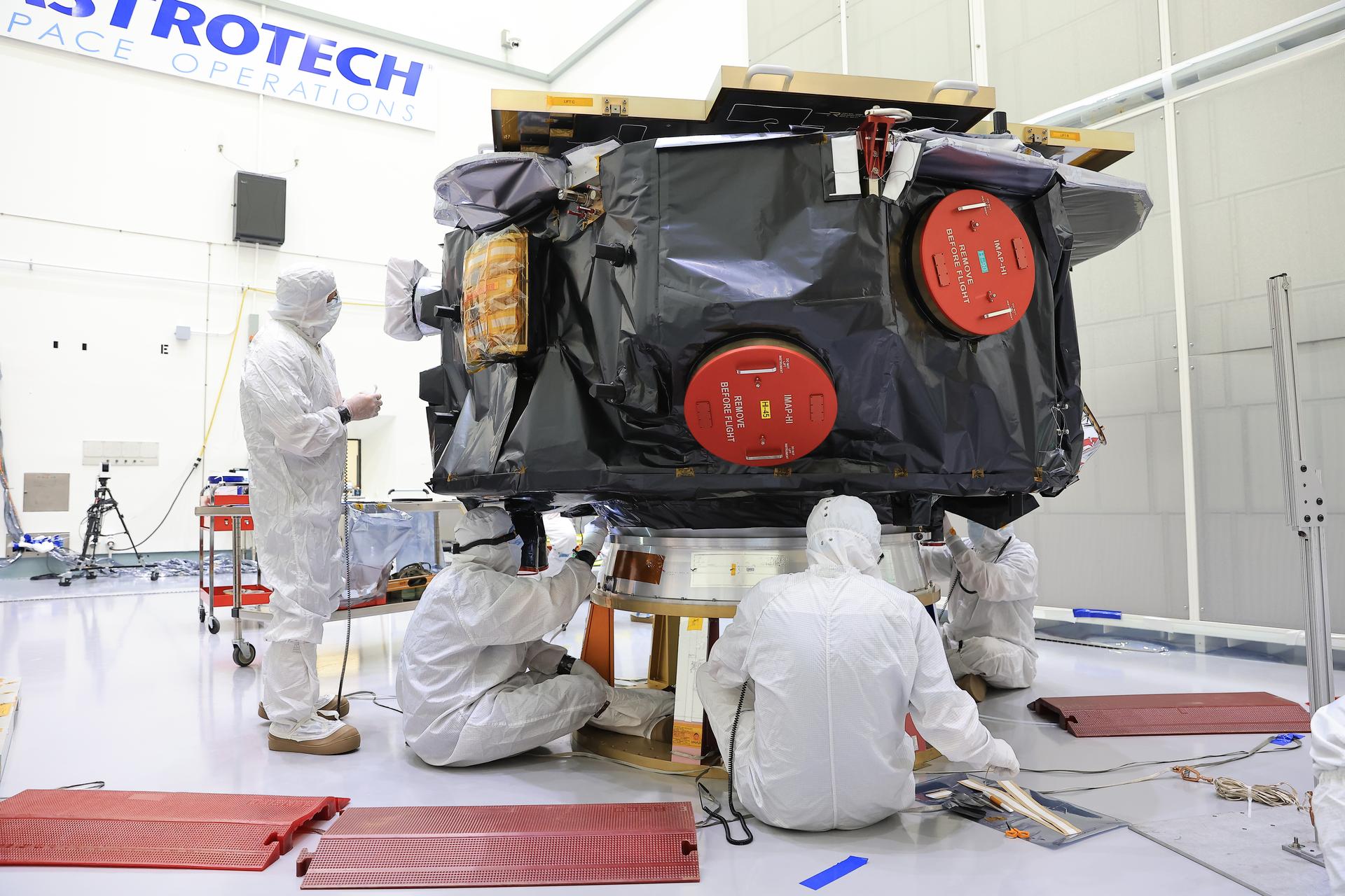 Image shows technicians in white cleanout suits, also known as bunny suits conducting blanket closeout work on NASA's IMAP mission, the black observatory on a stand.