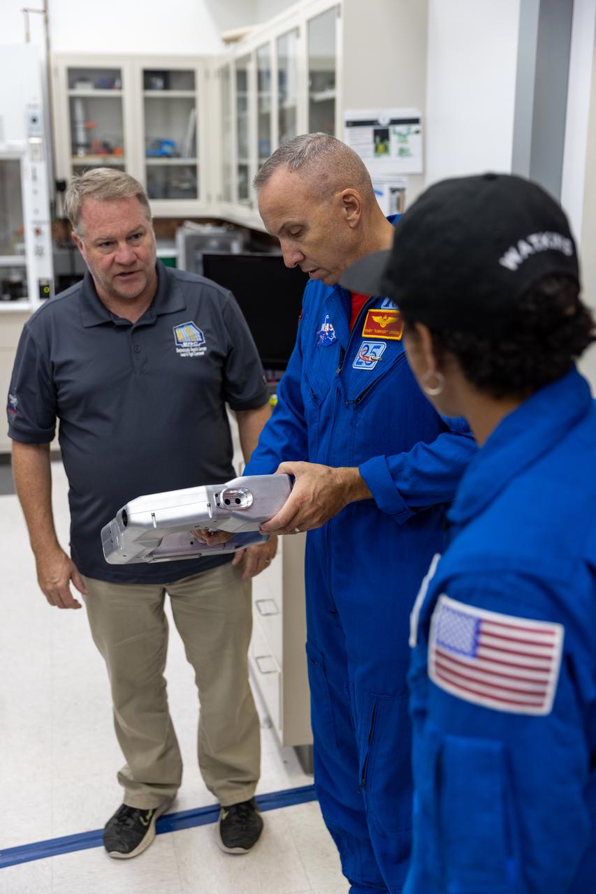 NASA astronauts Randy Bresnik (center) and Jessica Watkins (right) visit the Granular Mechanics and Regolith Operations Laboratory inside Swamp Works at Kennedy Space Center in Florida on Wednesday, Aug. 27, 2025, to view some of the evolving technologies in development that astronauts may use to explore the Moon’s surface, prepare it for sustainable outposts, and to handle the dust that is collected during moonwalks. Bresnick holds an instrument designed to help astronauts electromagnetically remove accumulated lunar dust.