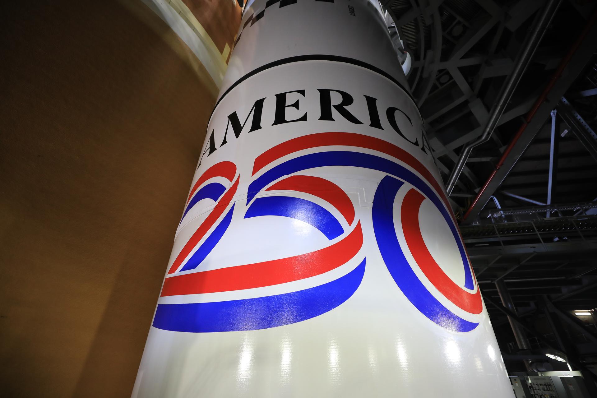 Technicians with NASA’s Exploration Ground Systems unveil on Monday, Dec. 1, 2025, the America 250 logo on the twin SLS (Space Launch System) solid rocket boosters for the Artemis II mission inside the Vehicle Assembly Building at NASA’s Kennedy Space Center in Florida. The SLS rocket and the Orion spacecraft will carry NASA astronauts Reid Wiseman, Victor Glover, Christina Koch, and CSA (Canadian Space Agency) astronaut Jeremy Hansen on a 10-day mission around the Moon and back in early 2026 from Launch Complex 39B at NASA Kennedy. America 250 commemorates the 250th anniversary of the signing of the Declaration of Independence with NASA celebrating the “Spirit of Innovation” theme to inspire future generations.