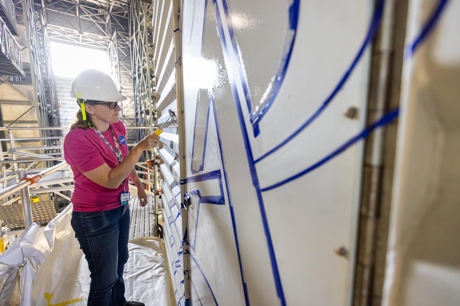 Katie Mortensen, mechanical engineering technician, paints NASA’s Artemis logo on the White Room connected to the crew access arm and mobile launcher inside the Vehicle Assembly Building at NASA’s Kennedy Space Center in Florida on Thursday, Dec. 4, 2025. The White Room is the area where the Artemis II crew Reid Wiseman, Victor Glover, Christina Koch, and CSA (Canadian Space Agency) astronaut Jeremy Hansen will enter the Orion spacecraft ahead of launch from Launch Complex 39B at NASA Kennedy in early 2026.