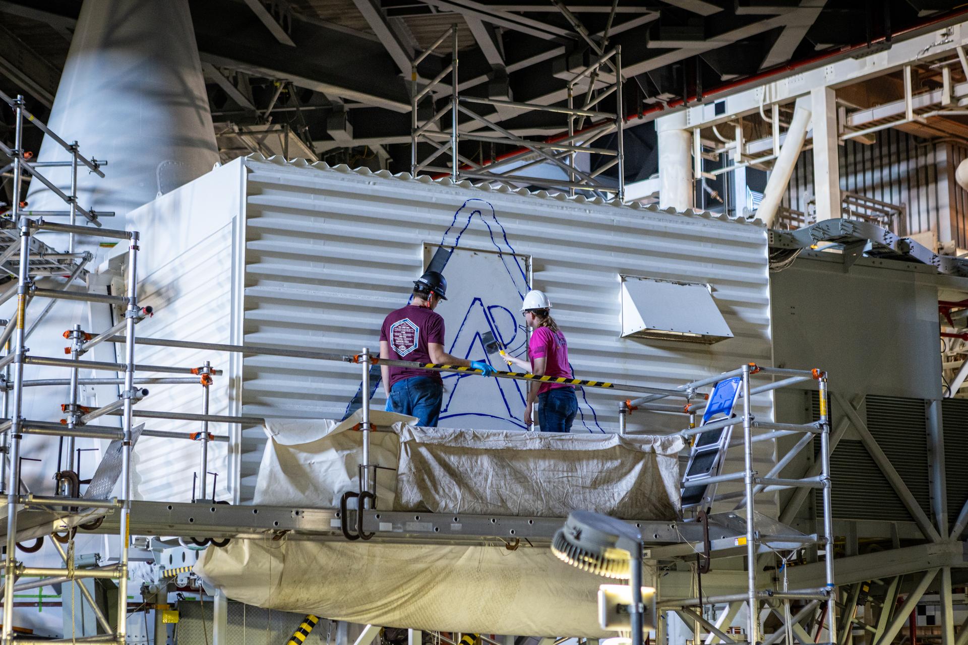 From left, Sean McCrary and Katie Mortensen, mechanical engineering technicians, paint NASA’s Artemis logo on the White Room connected to the crew access arm and mobile launcher inside the Vehicle Assembly Building at NASA’s Kennedy Space Center in Florida on Thursday, Dec. 4, 2025. The White Room is the area where the Artemis II crew Reid Wiseman, Victor Glover, Christina Koch, and CSA (Canadian Space Agency) astronaut Jeremy Hansen will enter the Orion spacecraft ahead of launch from Launch Complex 39B at NASA Kennedy in early 2026.