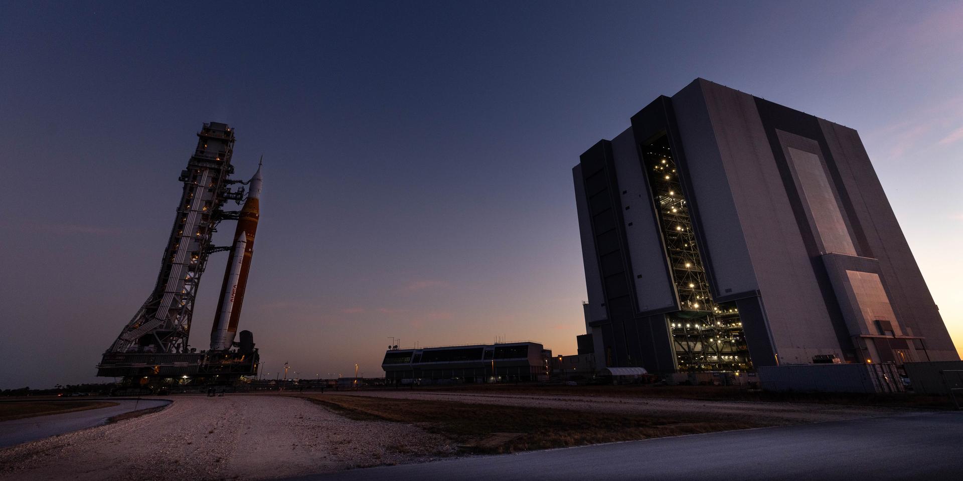 NASA’s crawler-transporter 2, carrying NASA’s Artemis II SLS (Space Launch System) rocket with the Orion spacecraft secured to mobile launcher 1, approaches the Vehicle Assembly Building at NASA’s Kennedy Space Center in Florida as the sun sets on Wednesday, Feb. 25, 2026, to troubleshoot the flow of helium to the rocket’s upper stage, the interim cryogenic propulsion stage. Seen in the background is also mobile launcher 2, which will be used on future Artemis flights beginning with Artemis IV. Once complete, the SLS rocket will roll back to Launch Complex 39B to prepare to launch four astronauts around the Moon and back for the Artemis II test flight. 