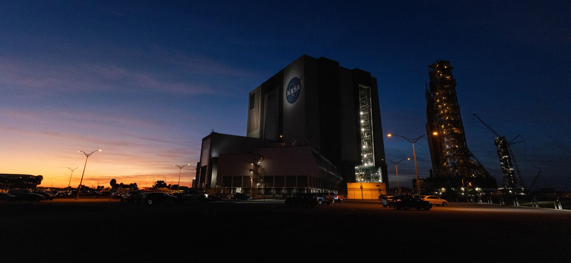NASA’s crawler-transporter 2, carrying NASA’s Artemis II SLS (Space Launch System) rocket with the Orion spacecraft secured to mobile launcher 1, approaches the Vehicle Assembly Building at NASA’s Kennedy Space Center in Florida as the sun sets on Wednesday, Feb. 25, 2026, to troubleshoot the flow of helium to the rocket’s upper stage, the interim cryogenic propulsion stage. Seen in the background is also mobile launcher 2, which will be used on future Artemis flights beginning with Artemis IV. Once complete, the SLS rocket will roll back to Launch Complex 39B to prepare to launch four astronauts around the Moon and back for the Artemis II test flight. 