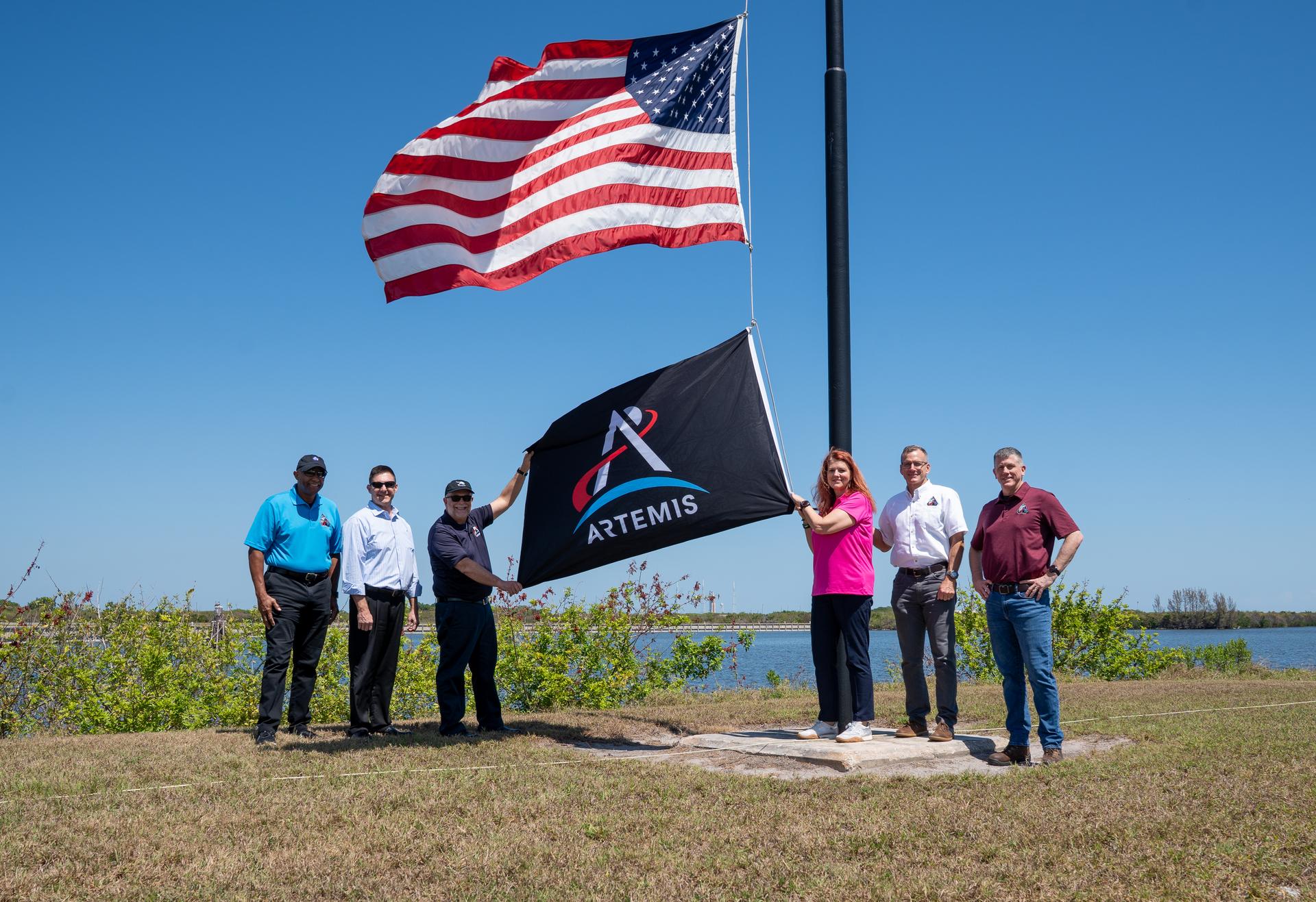 From left, NASA Kennedy Space Center Deputy Director Kelvin Manning; Robert Yaskovic, associate manager, Exploration Ground Systems Program; Shawn Quinn, manager, Exploration Ground Systems Program; Charlie Blackwell-Thompson, Artemis II launch director; Cliff Lanham, deputy manager, Exploration Ground Systems Program; and Jeremy Graeber, assistant launch director, Exploration Ground Systems Program raise the Artemis flag near the countdown clock at the NASA News Center at NASA’s Kennedy Space Center in Florida on Friday, March 27, 2026.  The Artemis II test flight mission will take Artemis II Commander Reid Wiseman, Pilot Victor Glover, and Mission Specialist Christina Koch from NASA, and Mission Specialist Jeremy Hansen from the CSA (Canadian Space Agency) on a 10-day journey around the Moon and back aboard NASA’s SLS (Space Launch System) rocket and Orion spacecraft from Launch Complex 39B no earlier than 6:24 p.m. EDT on Wednesday, April 1. 
