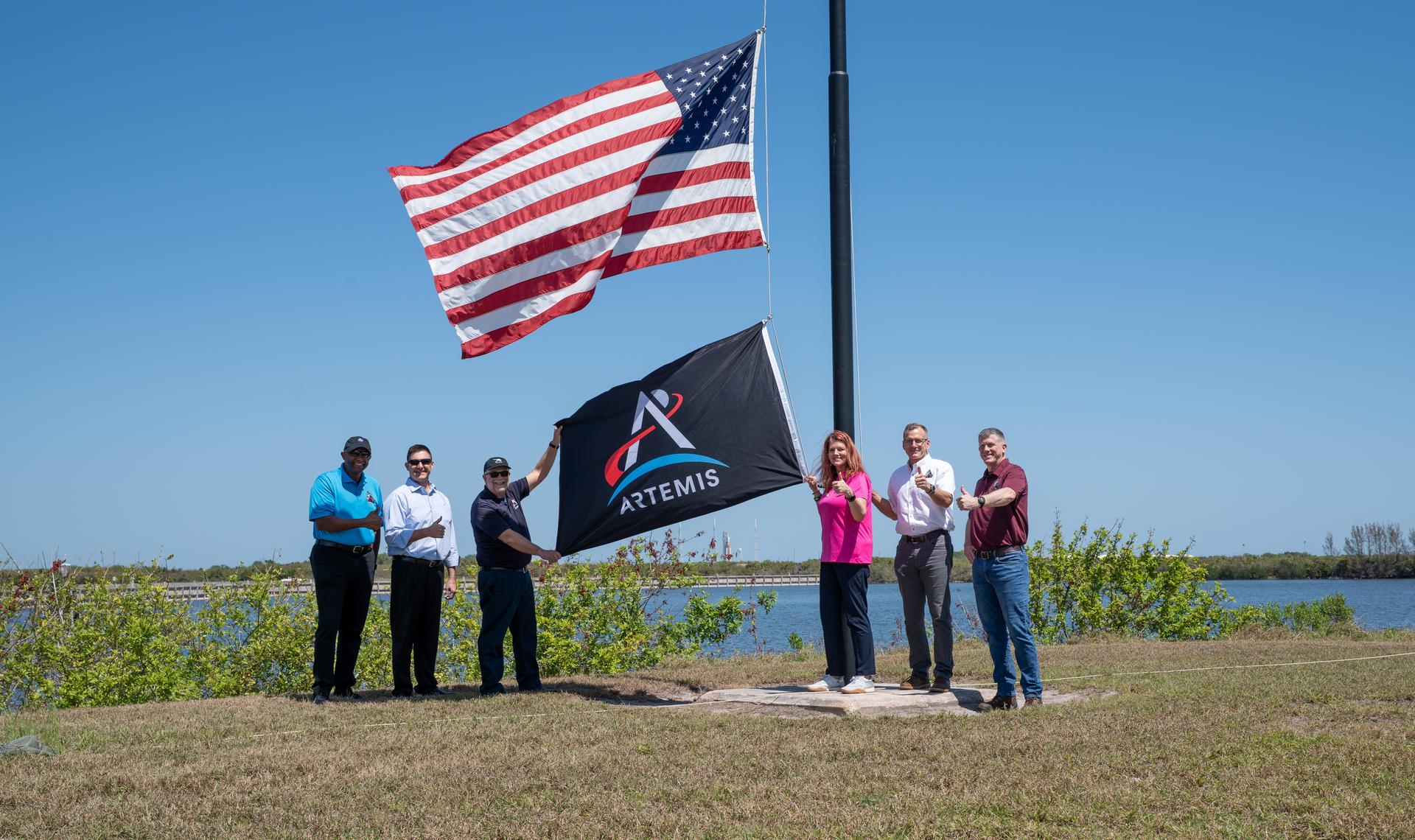 From left, NASA Kennedy Space Center Deputy Director Kelvin Manning; Robert Yaskovic, associate manager, Exploration Ground Systems Program; Shawn Quinn, manager, Exploration Ground Systems Program; Charlie Blackwell-Thompson, Artemis II launch director; Cliff Lanham, deputy manager, Exploration Ground Systems Program; and Jeremy Graeber, assistant launch director, Exploration Ground Systems Program raise the Artemis flag near the countdown clock at the NASA News Center at NASA’s Kennedy Space Center in Florida on Friday, March 27, 2026.  The Artemis II test flight mission will take Artemis II Commander Reid Wiseman, Pilot Victor Glover, and Mission Specialist Christina Koch from NASA, and Mission Specialist Jeremy Hansen from the CSA (Canadian Space Agency) on a 10-day journey around the Moon and back aboard NASA’s SLS (Space Launch System) rocket and Orion spacecraft from Launch Complex 39B no earlier than 6:24 p.m. EDT on Wednesday, April 1. 