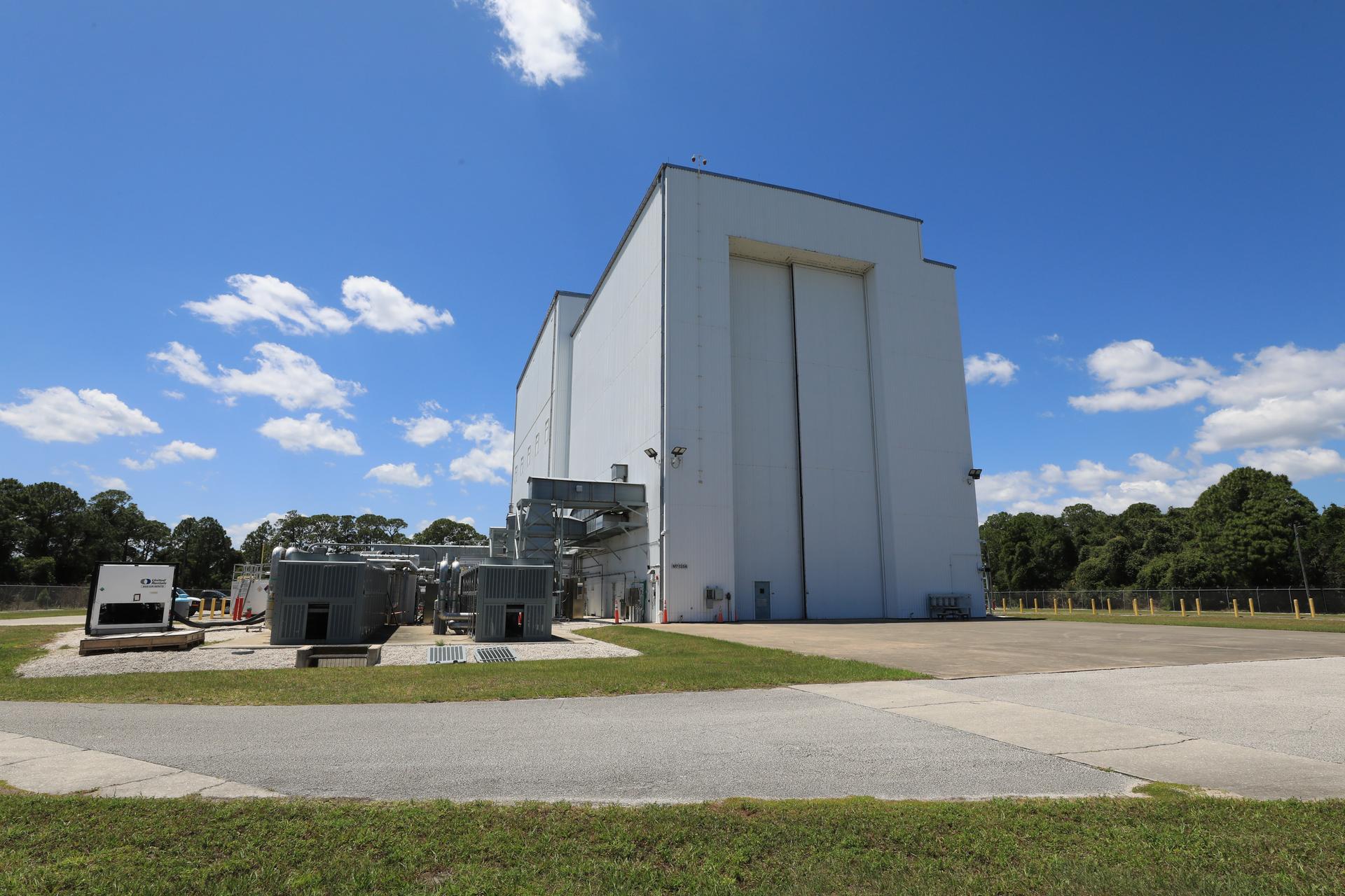 Image shows sunny, blue skies with a few wispy clouds outside the NASA Kennedy’s Payload Hazardous Servicing Facility (PHSF). The building is a grayish color with a nearby driveway and a small door for people to enter and huge doors for spacecrafts and instruments to enter. Photo credit: NASA/Kim Shiflett