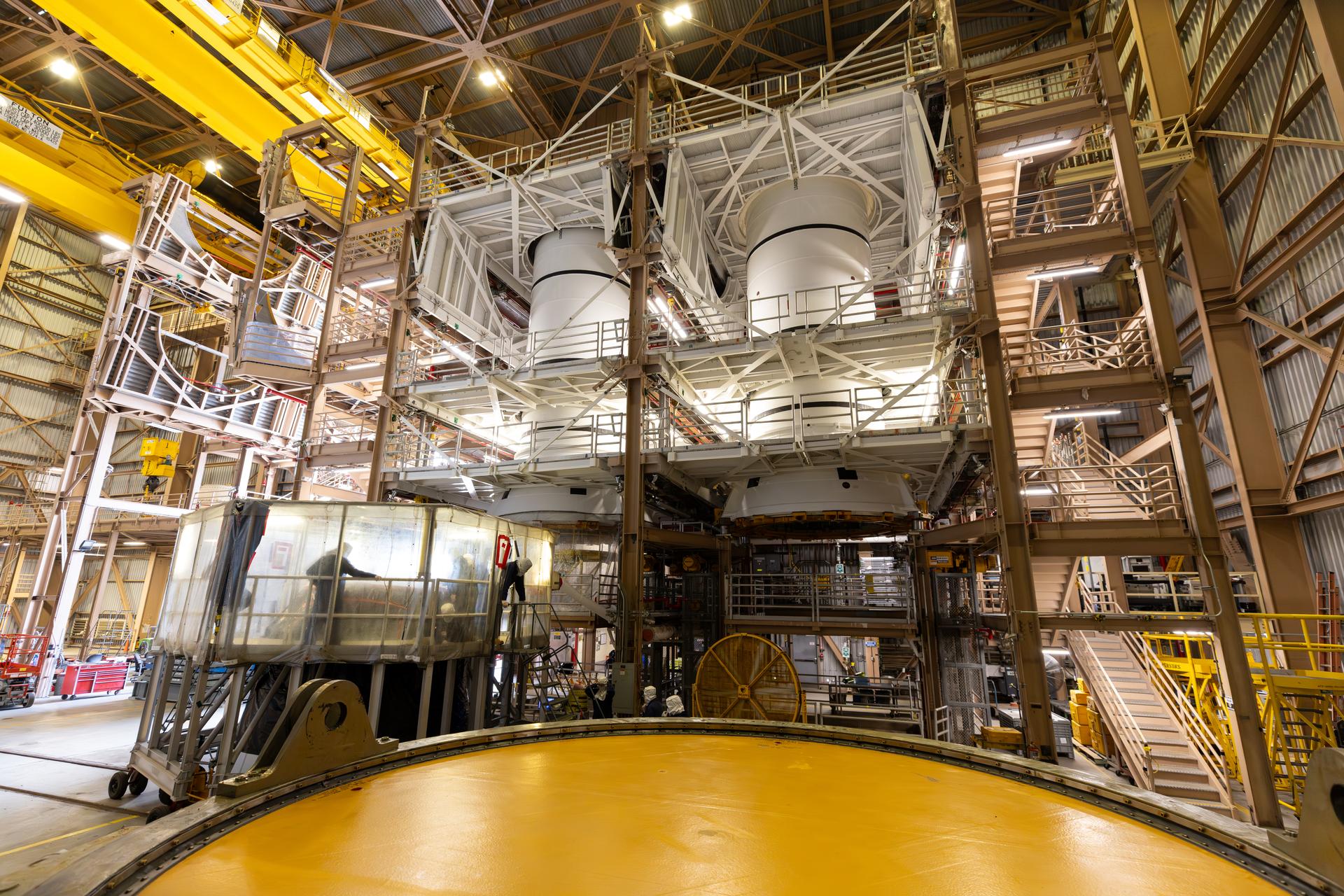 Inside a large industrial facility, two massive rocket engines or tank structures are suspended high within a metal scaffolding system. The surrounding area is filled with stairways, platforms, pipes, and support beams. In the foreground, a circular yellow platform dominates the lower part of the scene. Overhead lights illuminate the spacious interior, highlighting the complex machinery and structural framework.