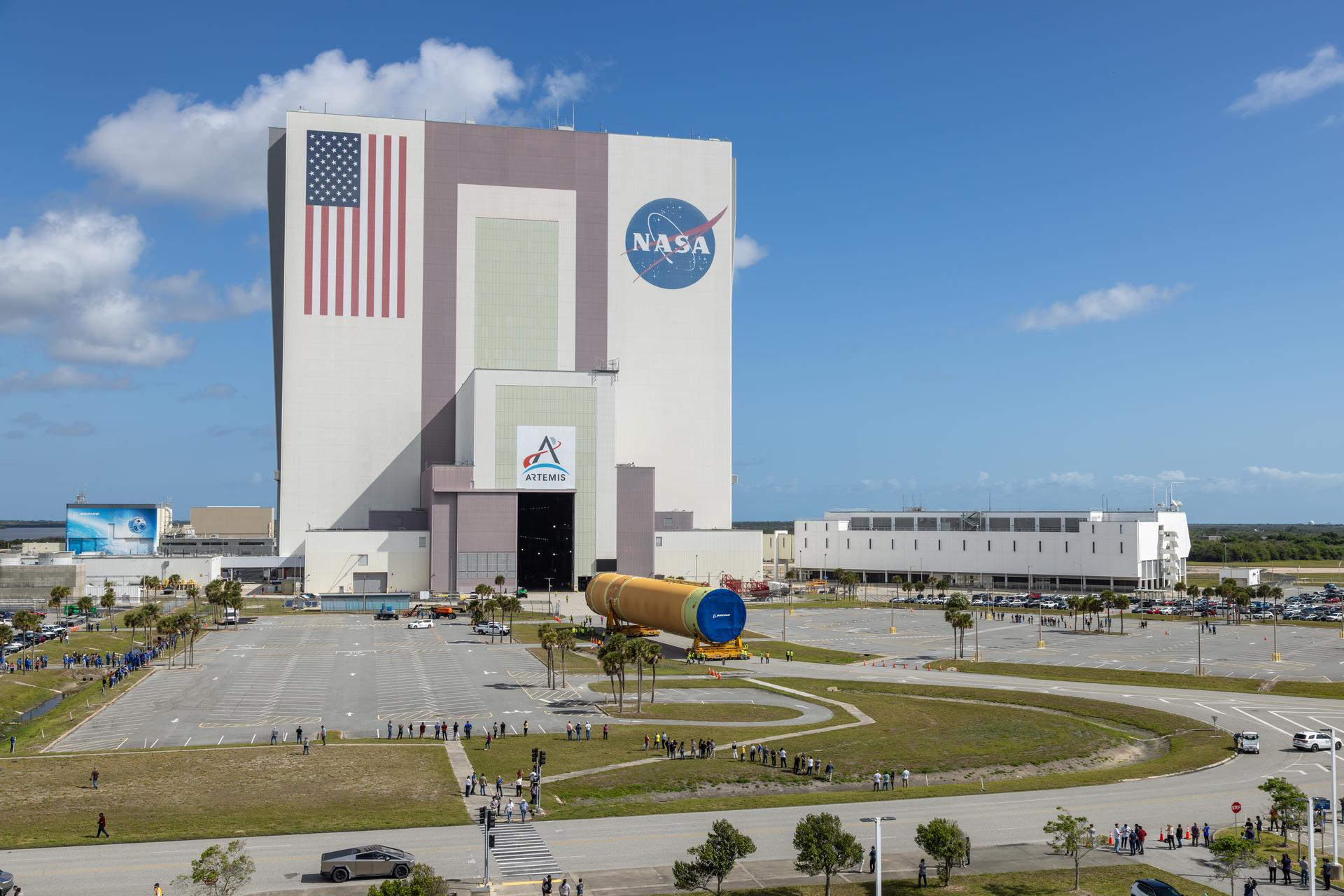 The image shows NASA's Kennedy Space Center iconic Vehicle Assembly Building with an American flag painted on the building along with the NASA and Artemis insignias in a blue sky with some clouds on Tuesday, April 28, 2026. People gathered to watch the transport of the Artemis III core stage, an orange yellow that will launch during the Artemis III mission. White buildings, grass, and a parking lot can also be seen in the photo. Photo credit: NASA/Glenn Benson