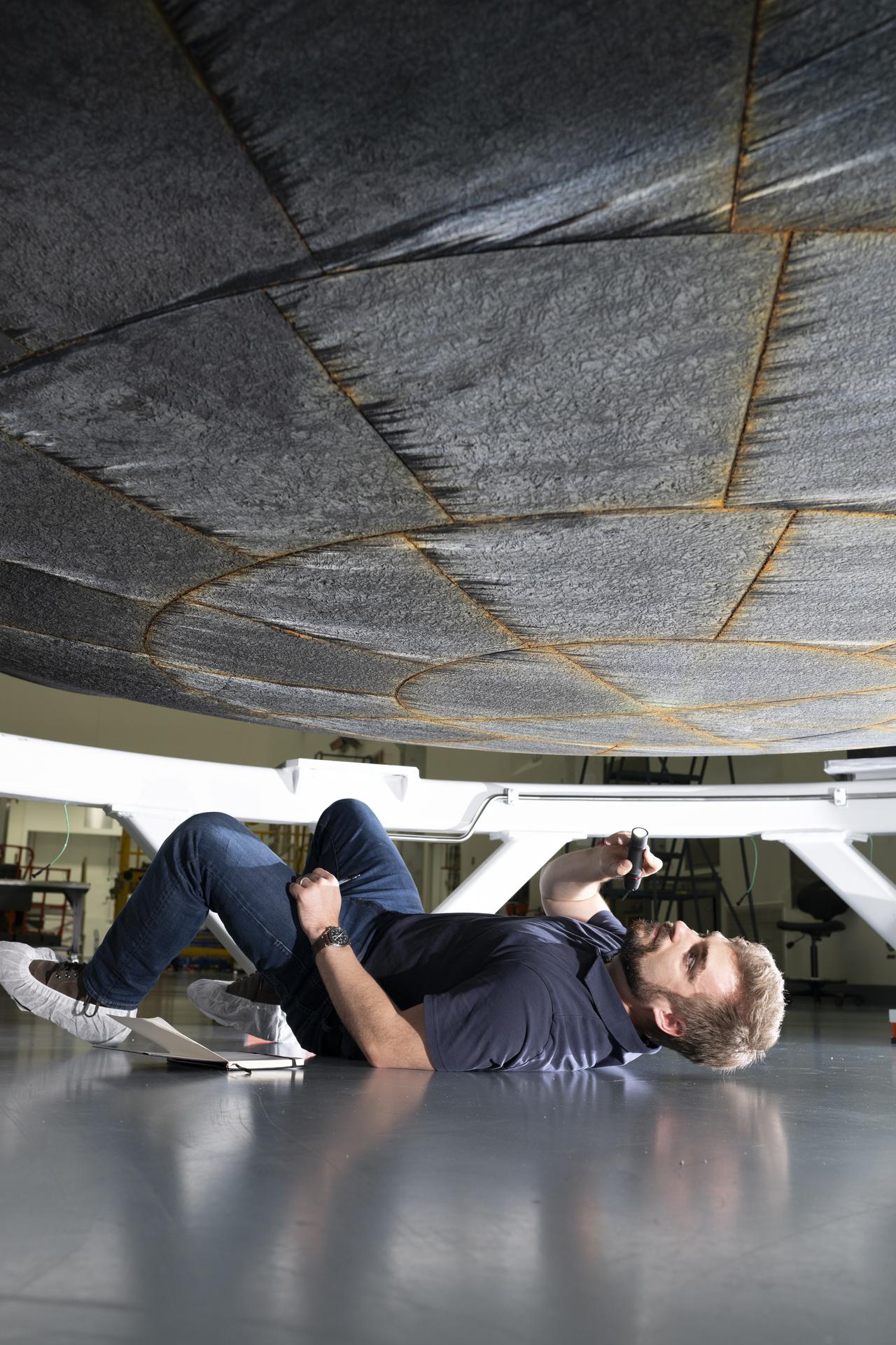 A person lies on the floor of a workshop, holding a flashlight to inspect the underside of a large, circular heat-shield-like structure suspended above them. The individual is positioned on their side beneath the hardware, with tools and equipment visible in the background.