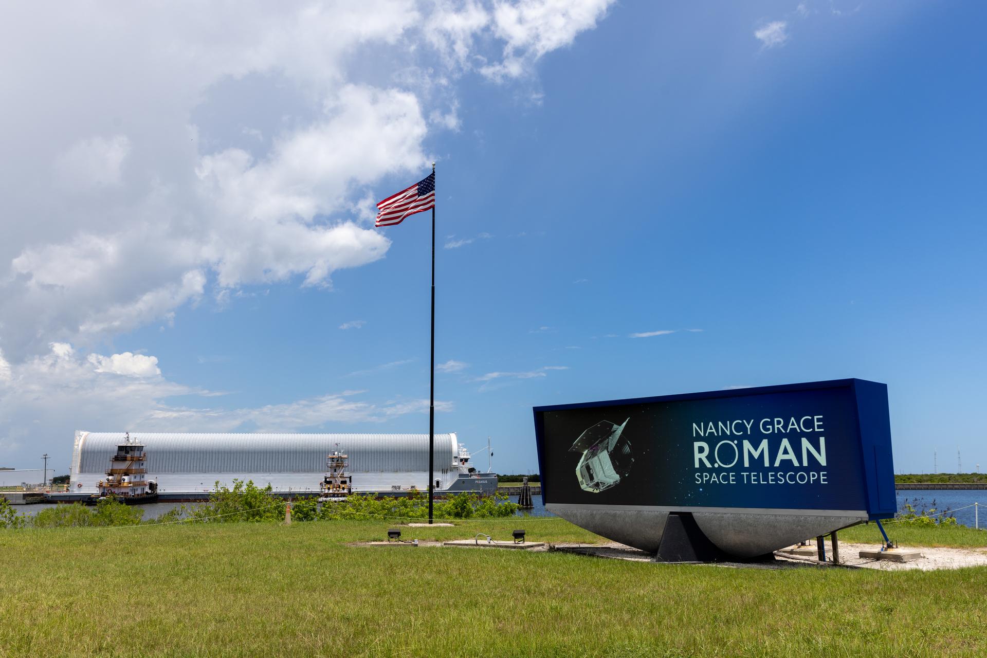 Image shows Nancy Grace Roman Telescope insignia on the countdown clock near an American flag on Sunday, June 21, 2026 at NASA's Kennedy Space Center in Florida. In the background is a white cylinder on a barge carrying NASA's Nancy Grace Roman Space Telescope with blue skies and a few puffy clouds. NASA/Amber Jean Notvest