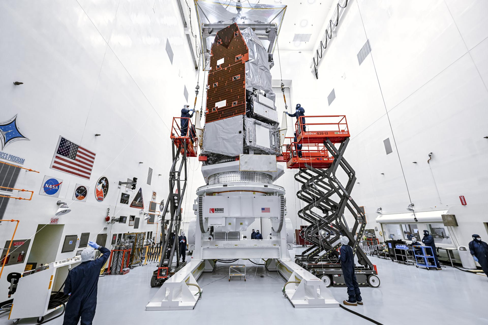 Technicians in clean room suits work around a tall spacecraft standing vertically on a large white support structure. Two scissor lifts on either side raise workers to access the spacecraft’s exterior, which is covered in silver insulation and copper-toned panels. The bright white room features mission logos and flags on the wall, with equipment and workstations arranged around the space. Photo credit: NASA/Sydney Rohde (Rocz)