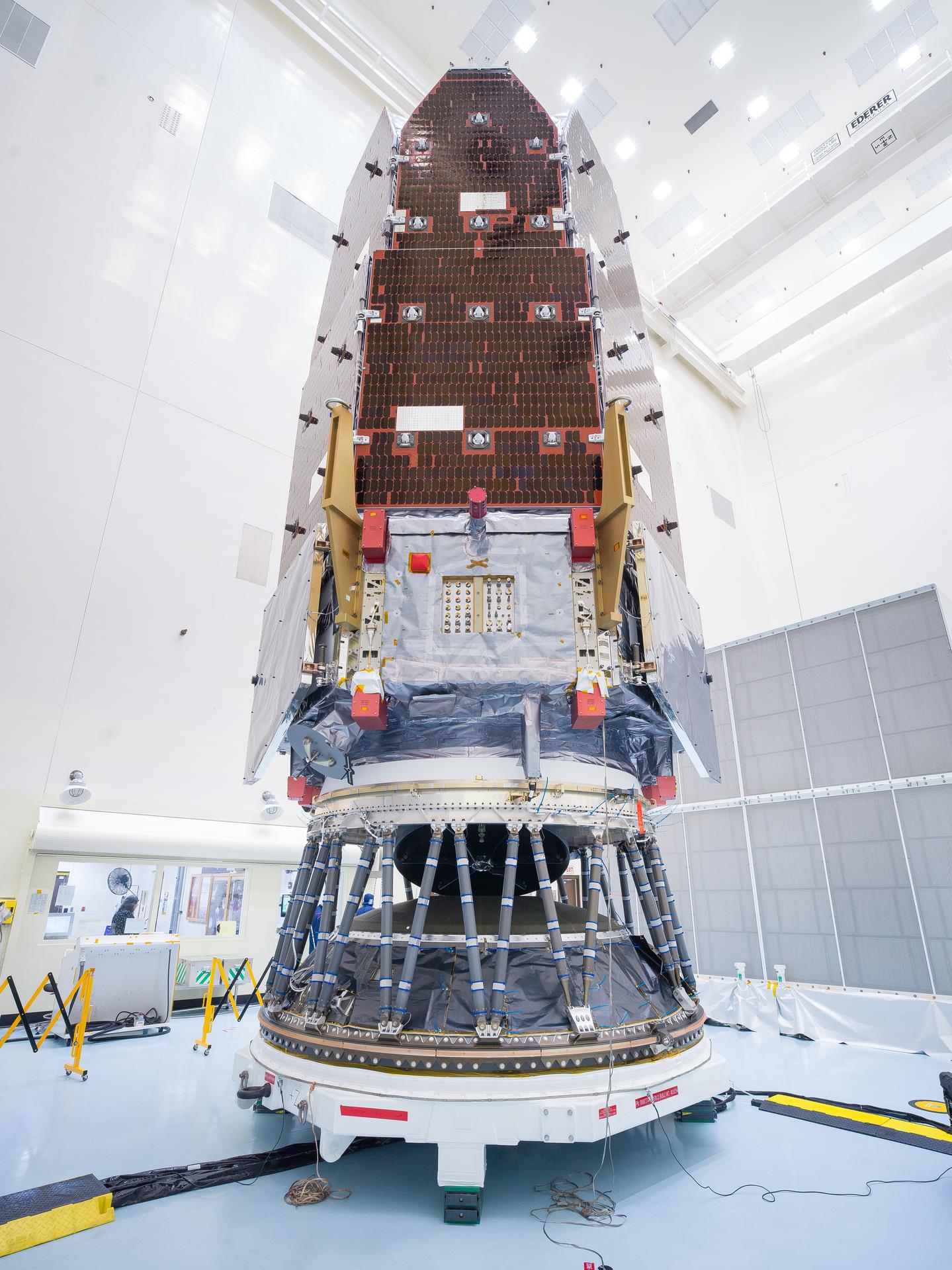 Image shows a white clean room at Kennedy Space Center in Florida. A large metallic looking spacecraft is vertical on a cyclinder piece of hardware that is secured to a workstand with wheels. In the middle of the spacecraft is red, orange colored solar array panels.