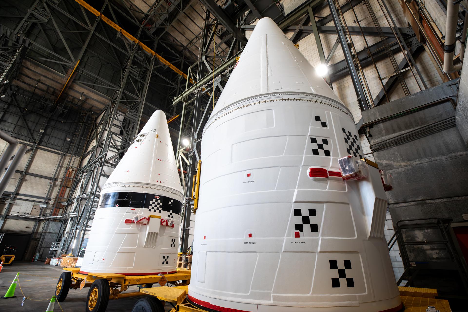 The left, with black stripe, and right aerodynamic forward assemblies for the twin SLS (Space Launch System) solid rocket boosters for Artemis III arrive at the Vehicle Assembly Building at NASA’s Kennedy Space Center in Florida on Thursday, Aug. 13, 2026. The nose cones will be integrated atop the SLS boosters to provide a smooth, pointed shape that allows the boosters to cut through the atmosphere efficiently, reducing drag and keeping the rocket stable during ascent. Artemis III will carry out a series of objectives in low Earth orbit designed to demonstrate critical systems needed for future lunar landings, beginning with Artemis IV.  