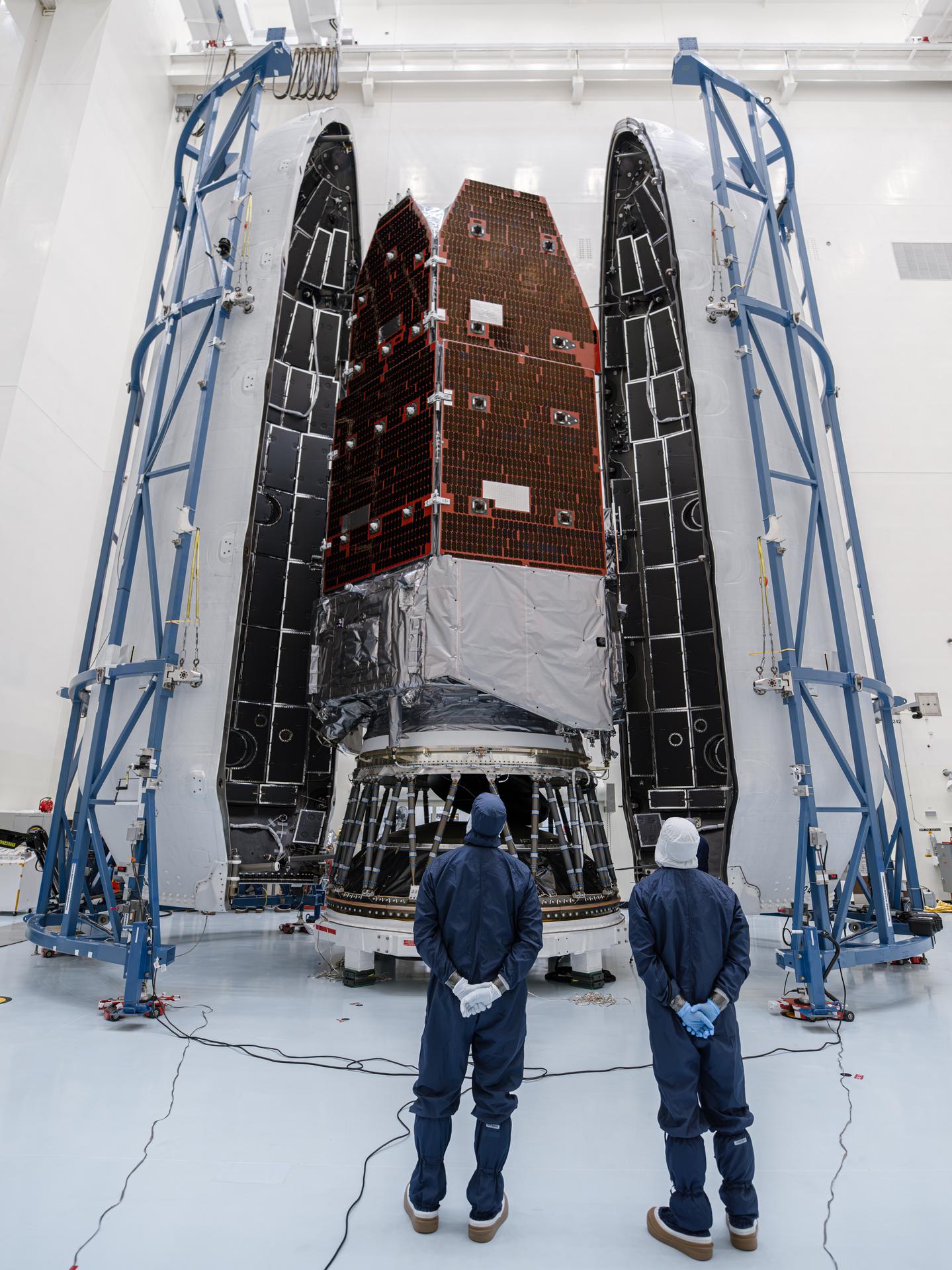Two technicians in cleanroom suits stand in front of the Roman Space Telescope and payload fairing, which tower above them inside the high-bay facility.
