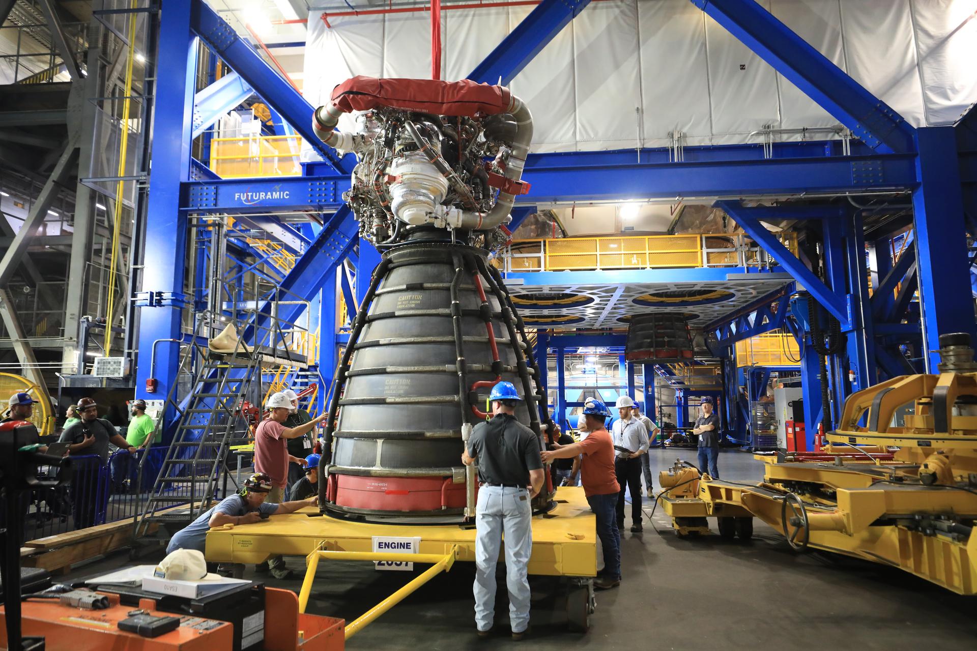 A rocket engine sits on a yellow transport platform inside a large industrial facility. Several technicians wearing hard hats and safety gear stand around the engine, inspecting and guiding it. The engine’s bell nozzle faces downward, and complex piping and hardware are visible at the top. Bright blue structural frames and elevated yellow walkways surround the work area, with additional equipment and machinery in the background.